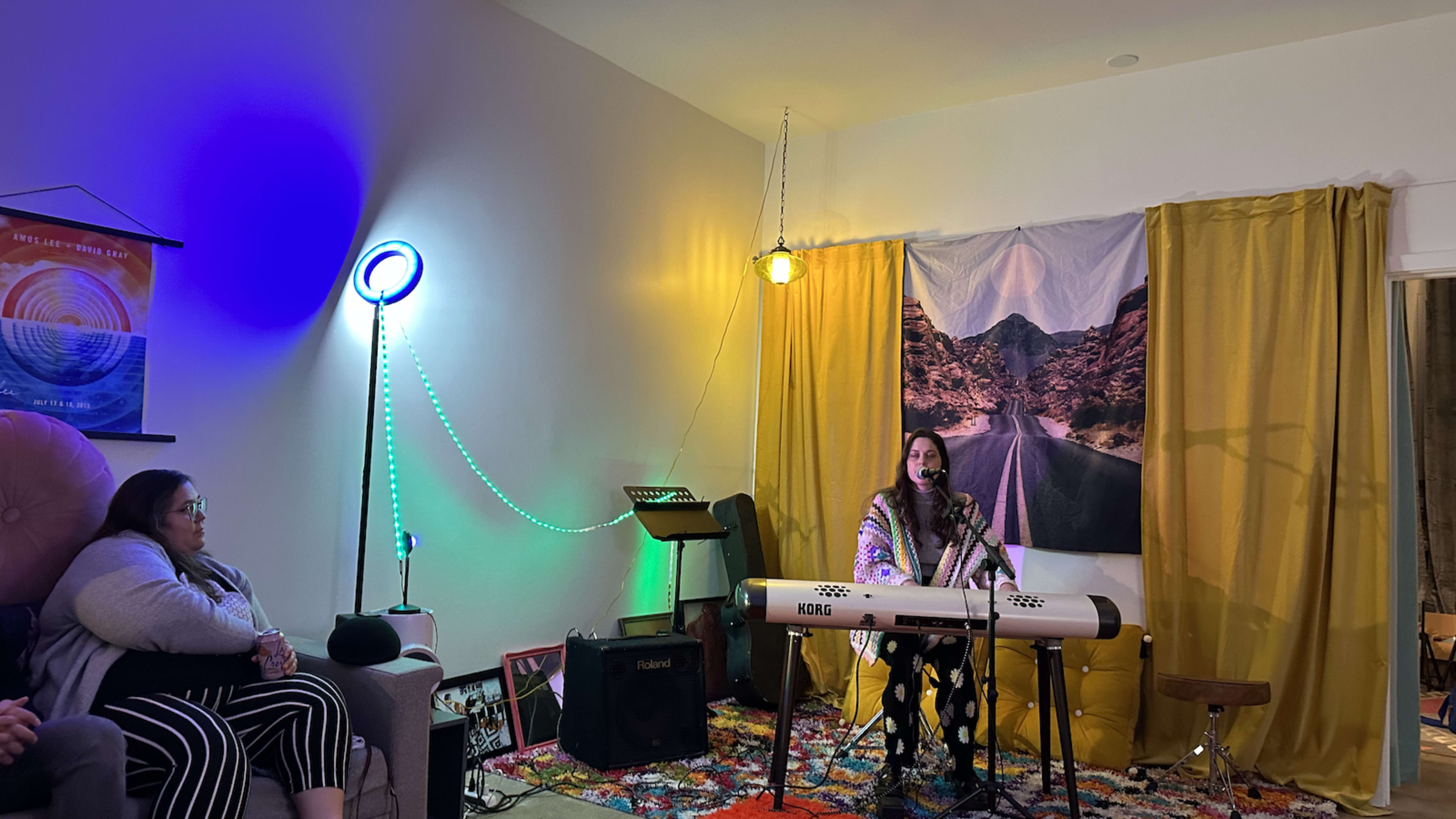 A woman sings and plays a keyboard in a cozy, colorful room while two audience members watch.