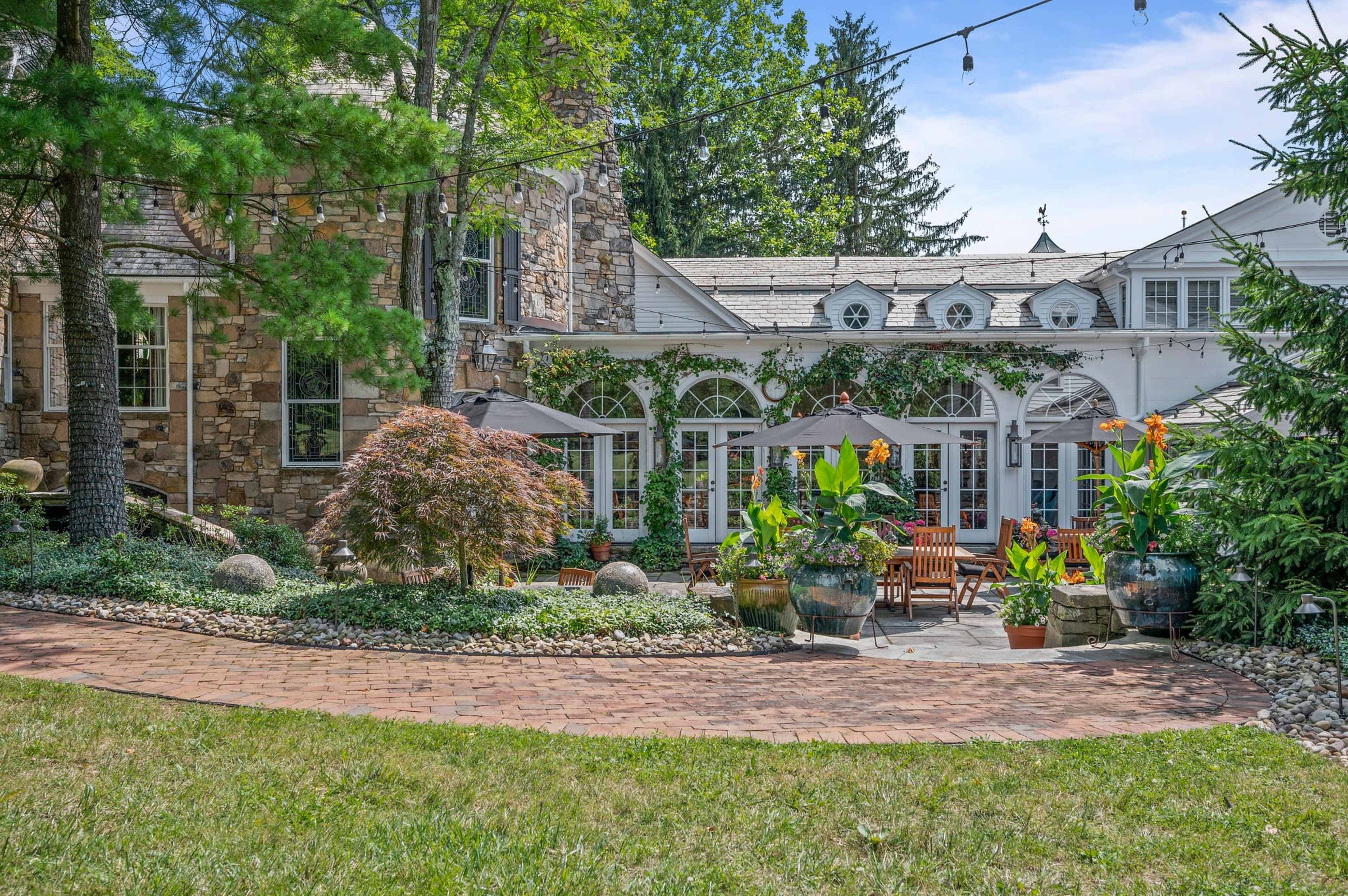 The image shows a landscaped outdoor area of a stone-clad house featuring a patio with seating, potted plants, and string lights.