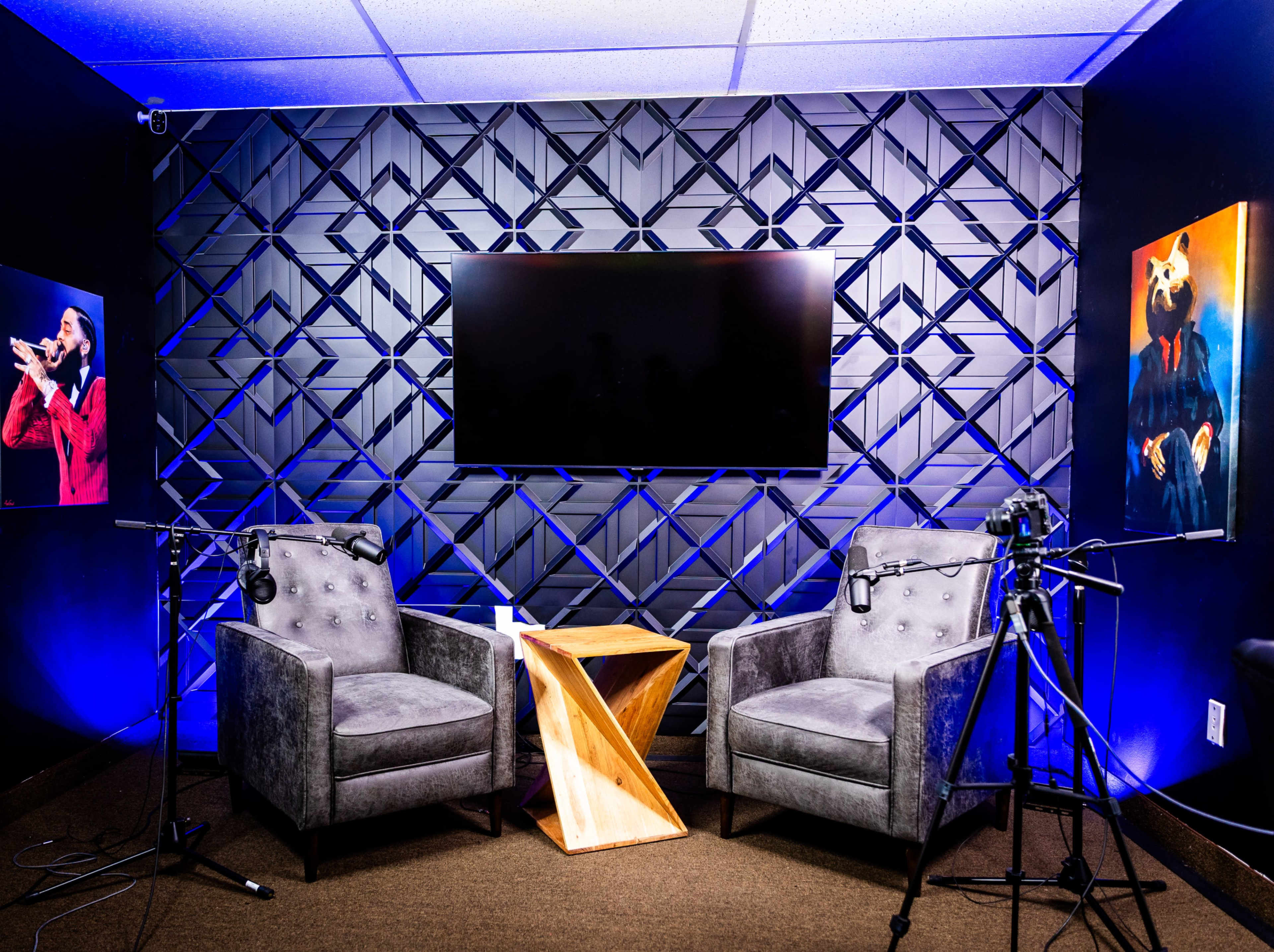The image shows a modern recording studio setup with two upholstered chairs, a wooden side table, and a wall-mounted screen surrounded by artistic wall decor and microphones.