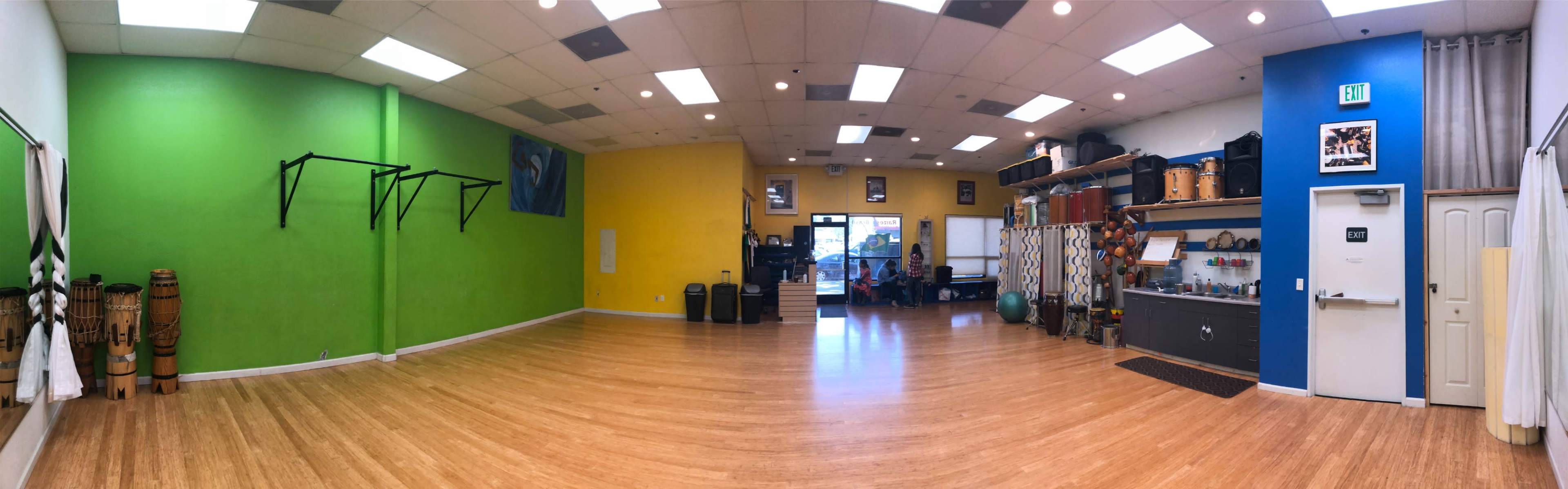 A spacious interior of a studio with a green and yellow wall, wooden flooring, various exercise equipment, and people engaged in activities near the entrance.