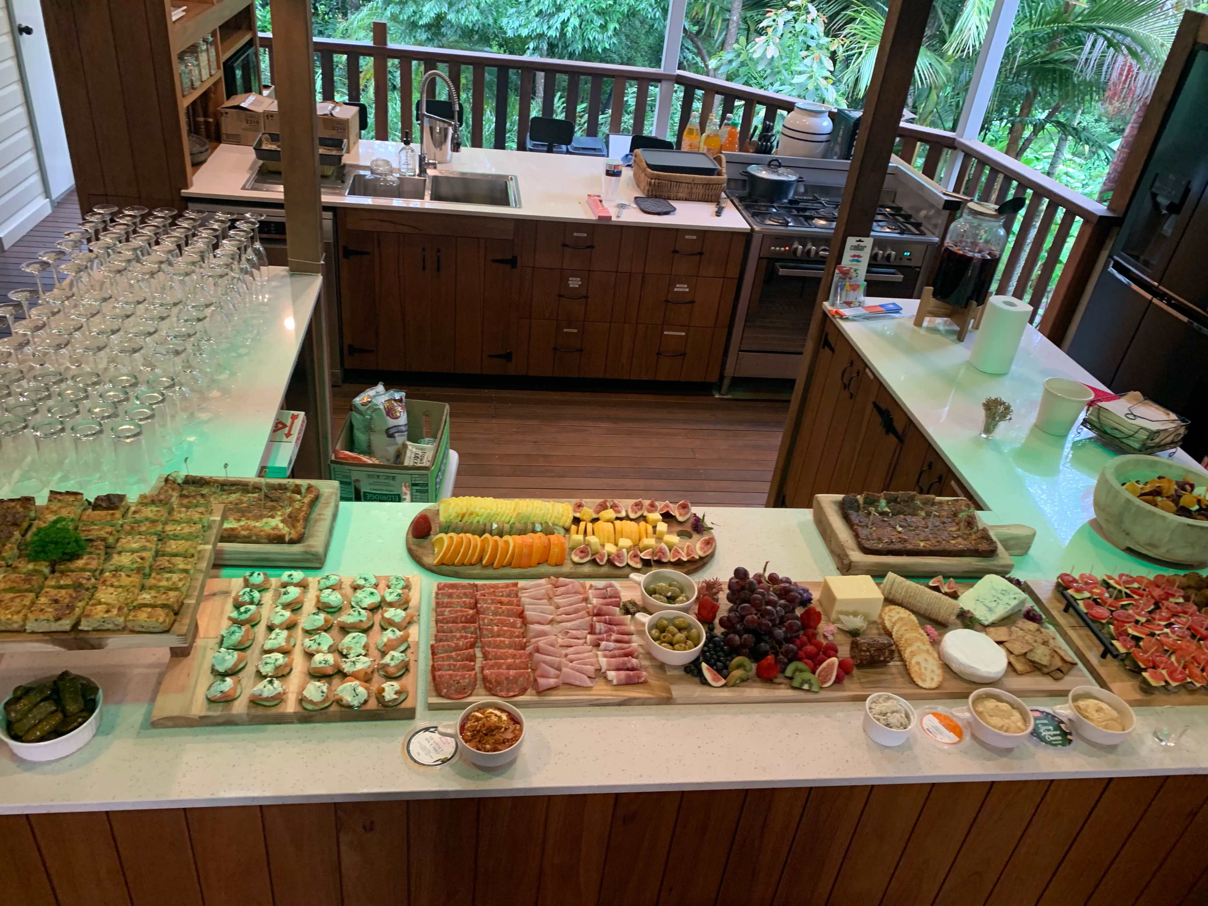 A large spread of food, including various fruits, appetizers, and desserts, is arranged on a countertop in a kitchen surrounded by greenery.