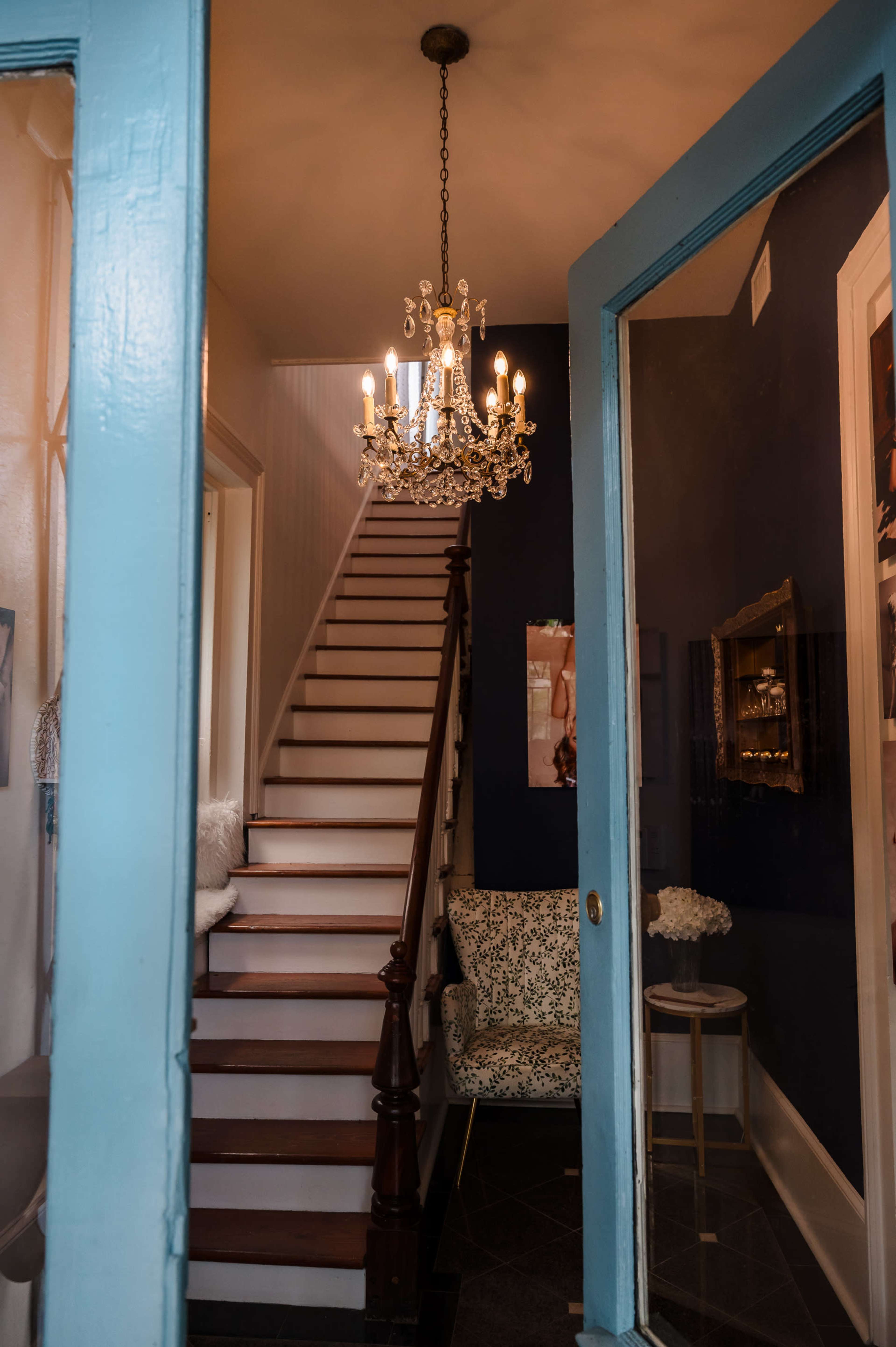 A bright entryway features a staircase leading upward, adorned with a chandelier above and a patterned chair to the side.