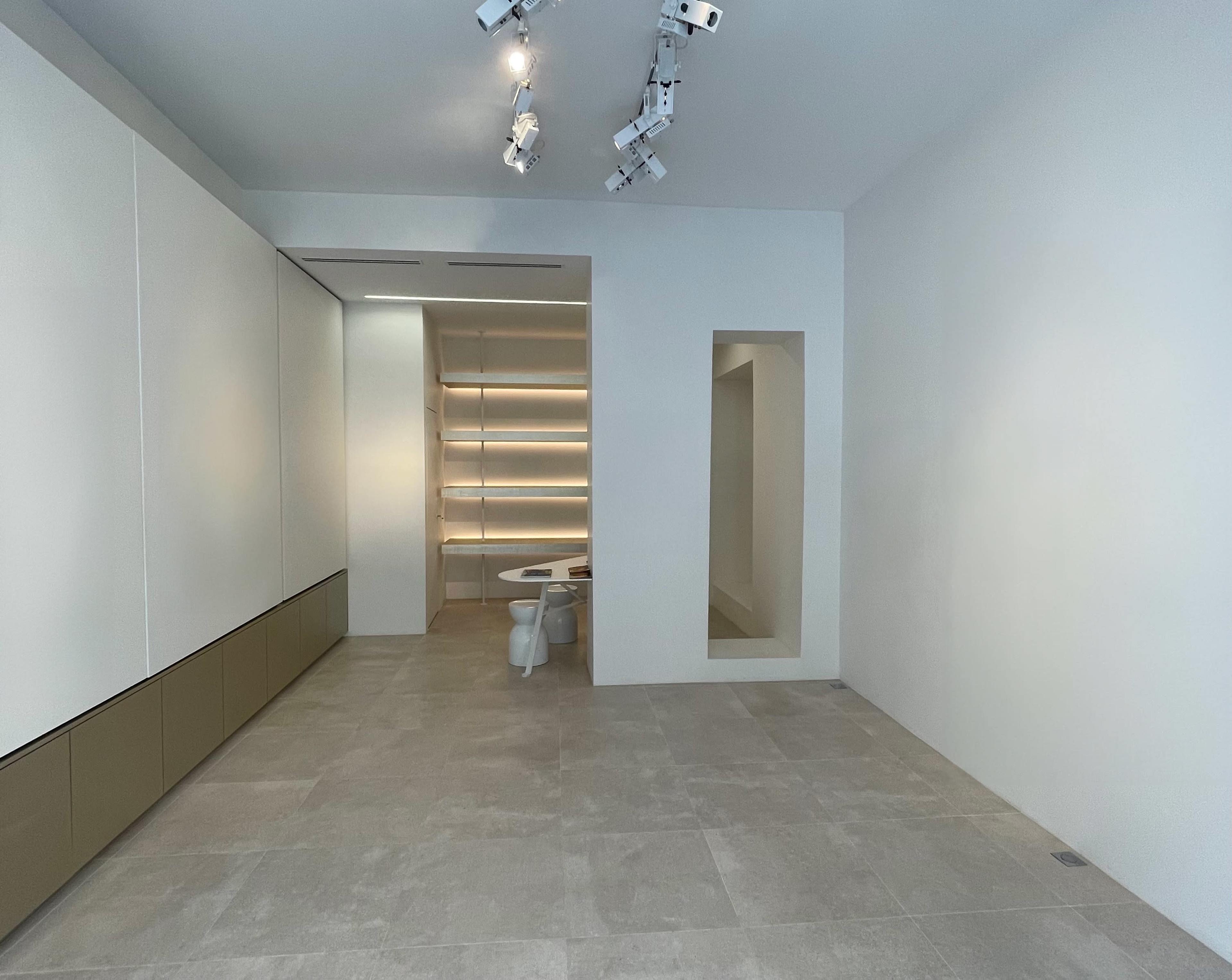 A minimalist interior space featuring light-colored walls, tiled flooring, built-in shelving with soft lighting, and a circular table with chairs.