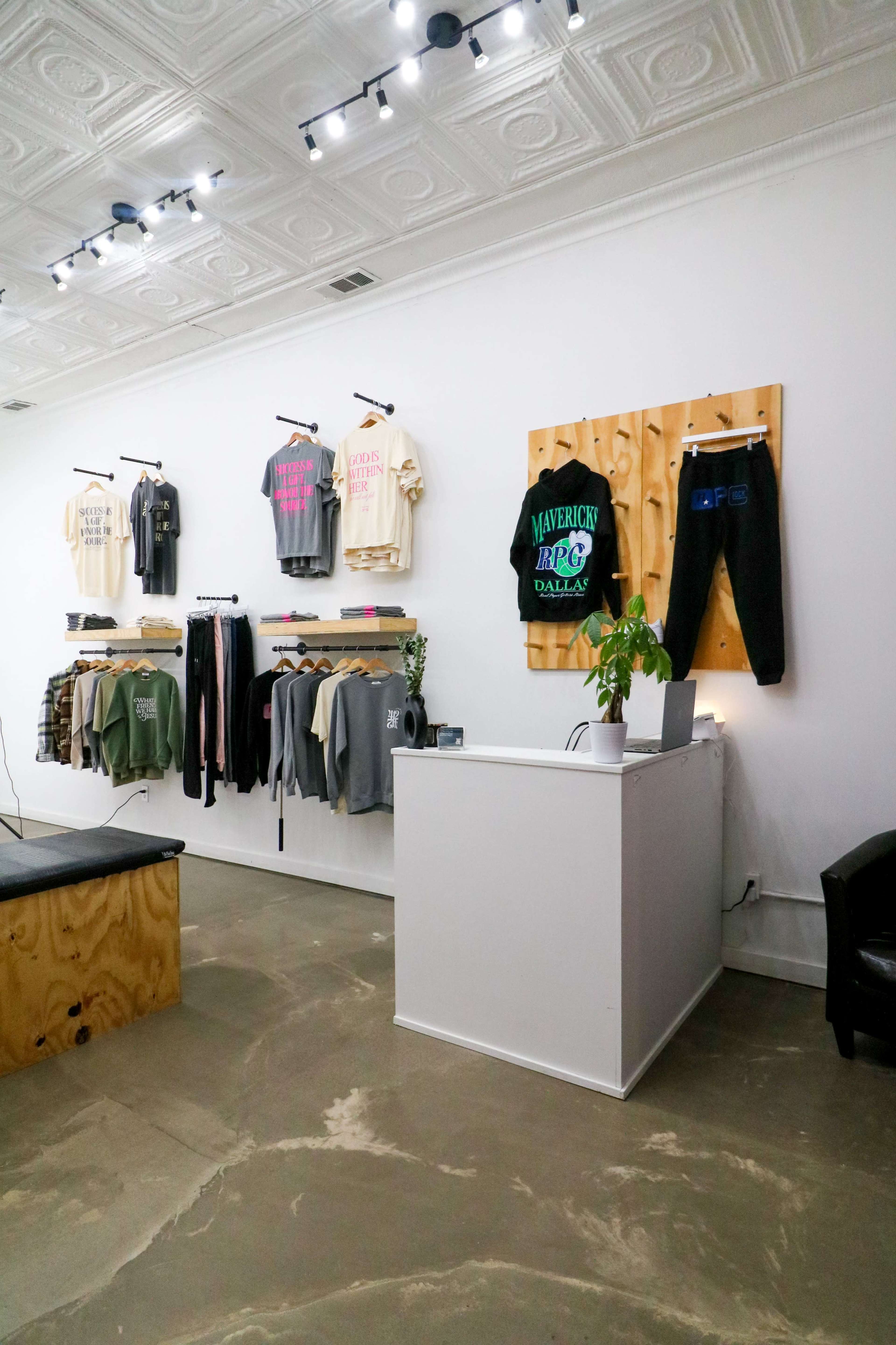 The image shows a modern retail space with clothing hung on the walls and a counter at the front with a plant and a computer.