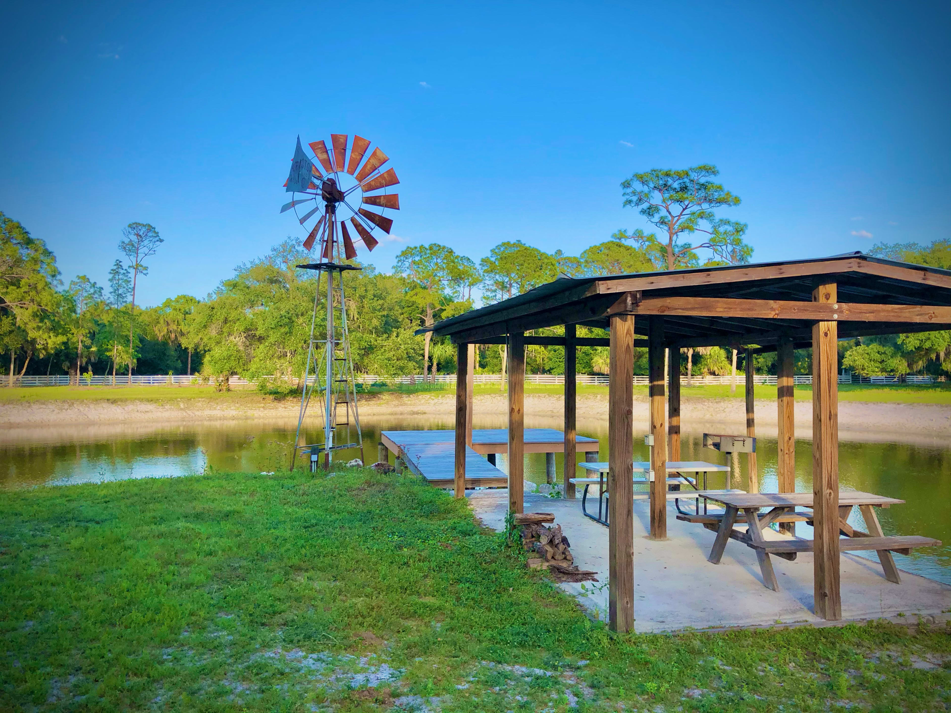 A wooden gazebo with picnic tables is positioned near a calm pond, alongside a tall windmill.