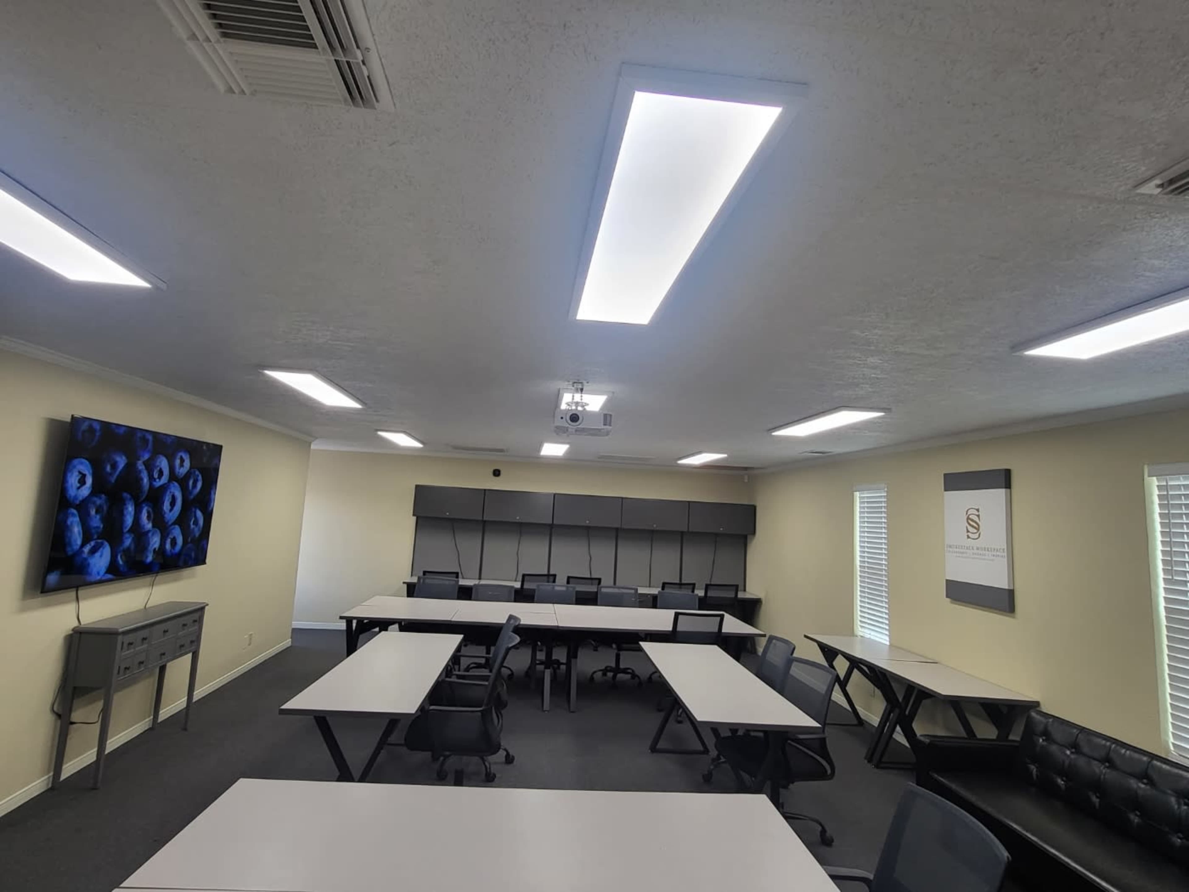 The image shows a modern conference room with several rectangular tables arranged in a U-shape, black chairs, a projector on the ceiling, and a screen displaying an image of blueberries on the wall.