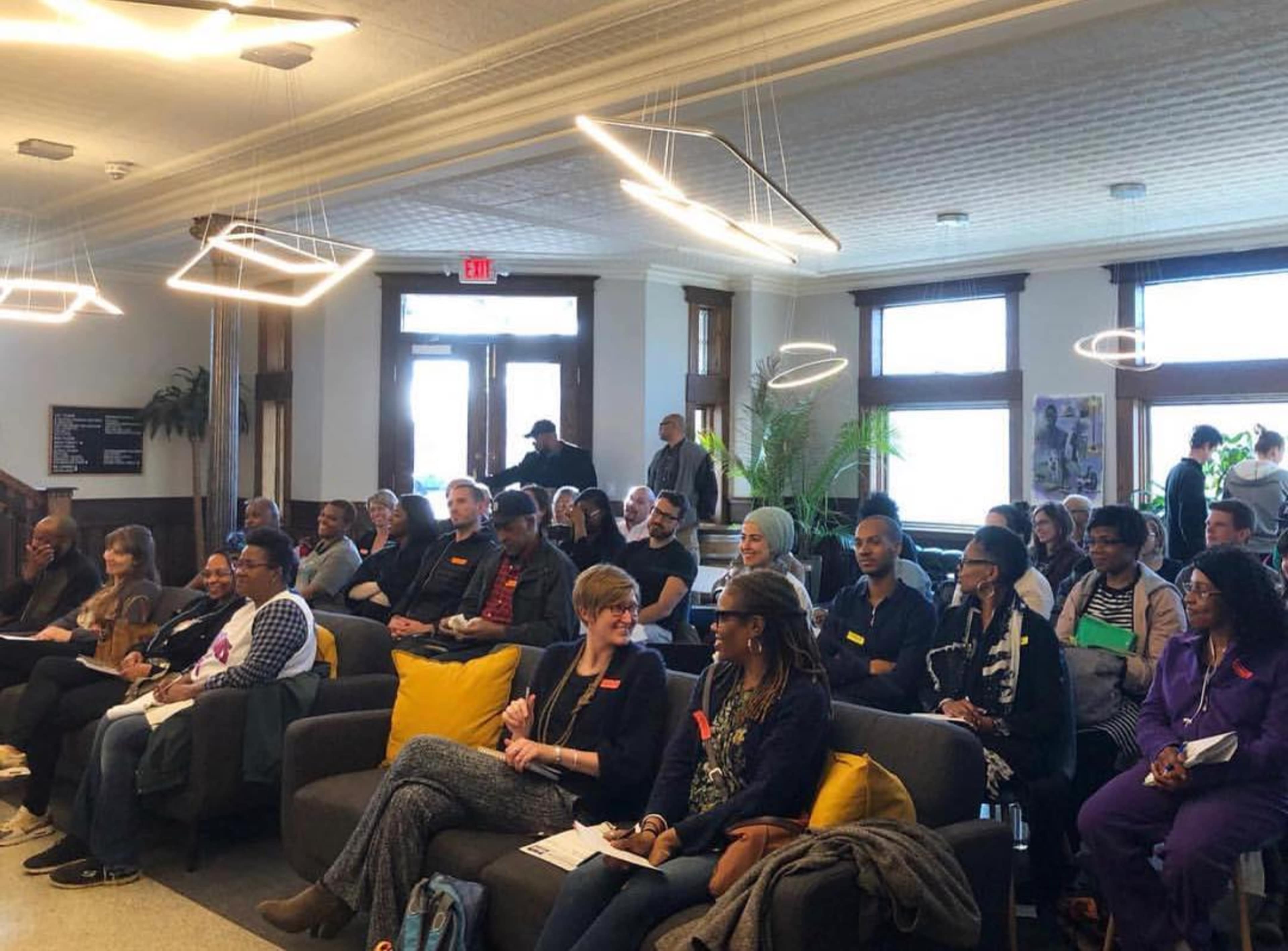 A group of people sits in a modern lounge with large windows and comfortable seating, attending an event.