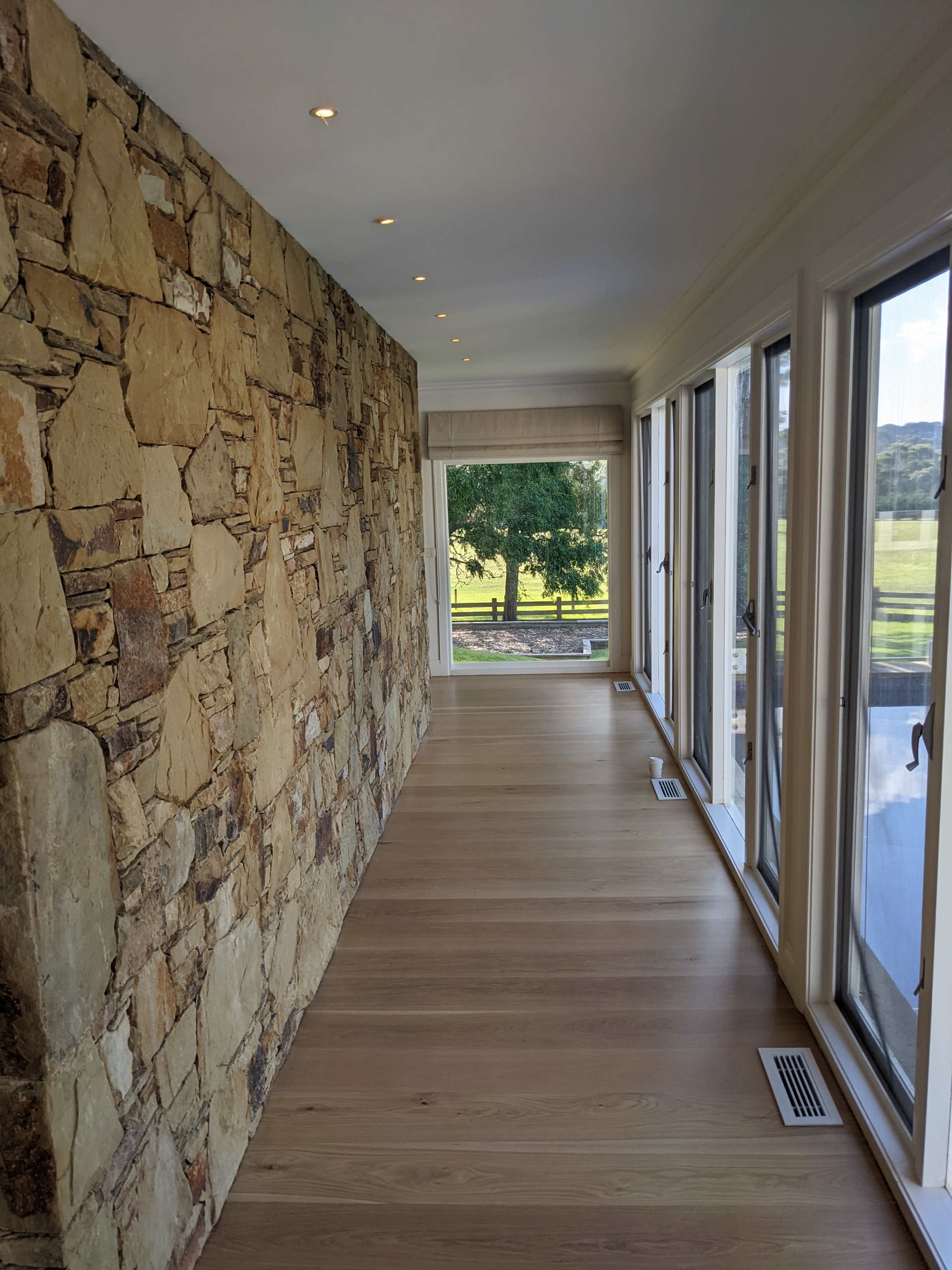 The image shows a narrow hallway featuring a stone wall on one side and large glass windows on the opposite side, leading to a view of greenery outside.