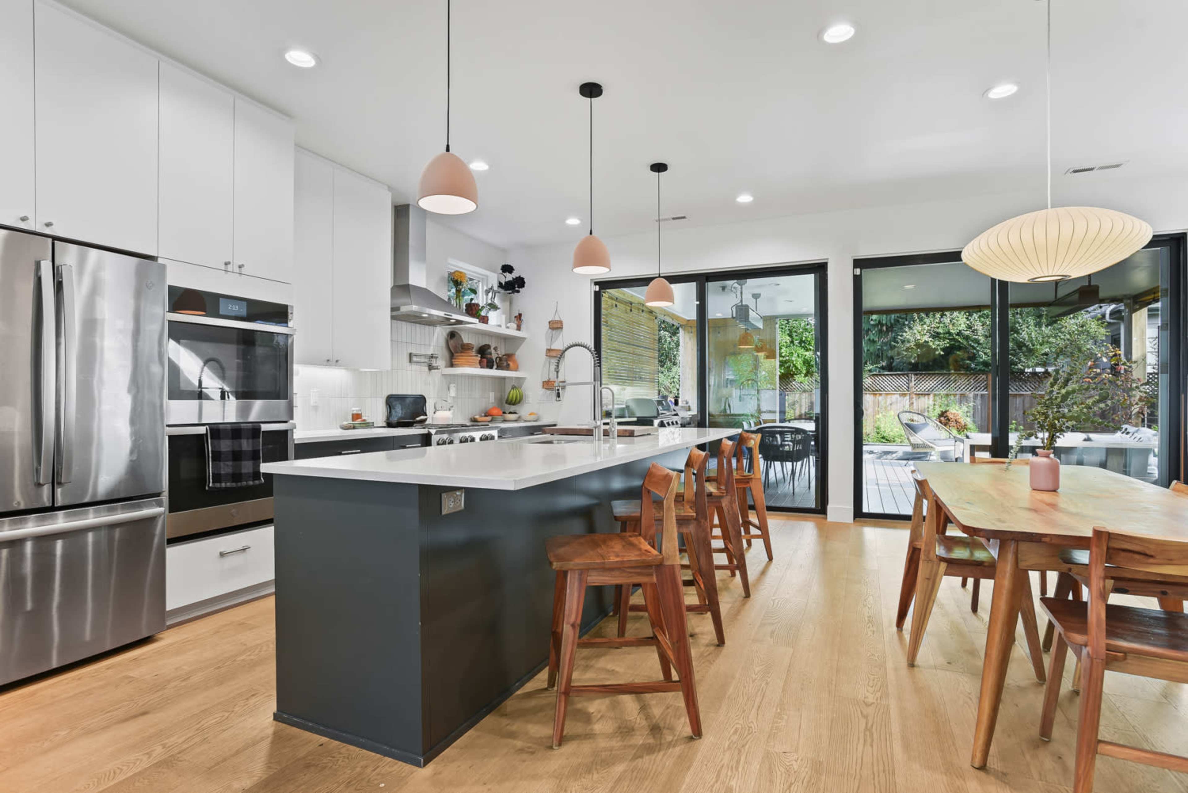 A modern kitchen features a large island with bar stools, sleek appliances, and ample natural light from windows facing a patio.