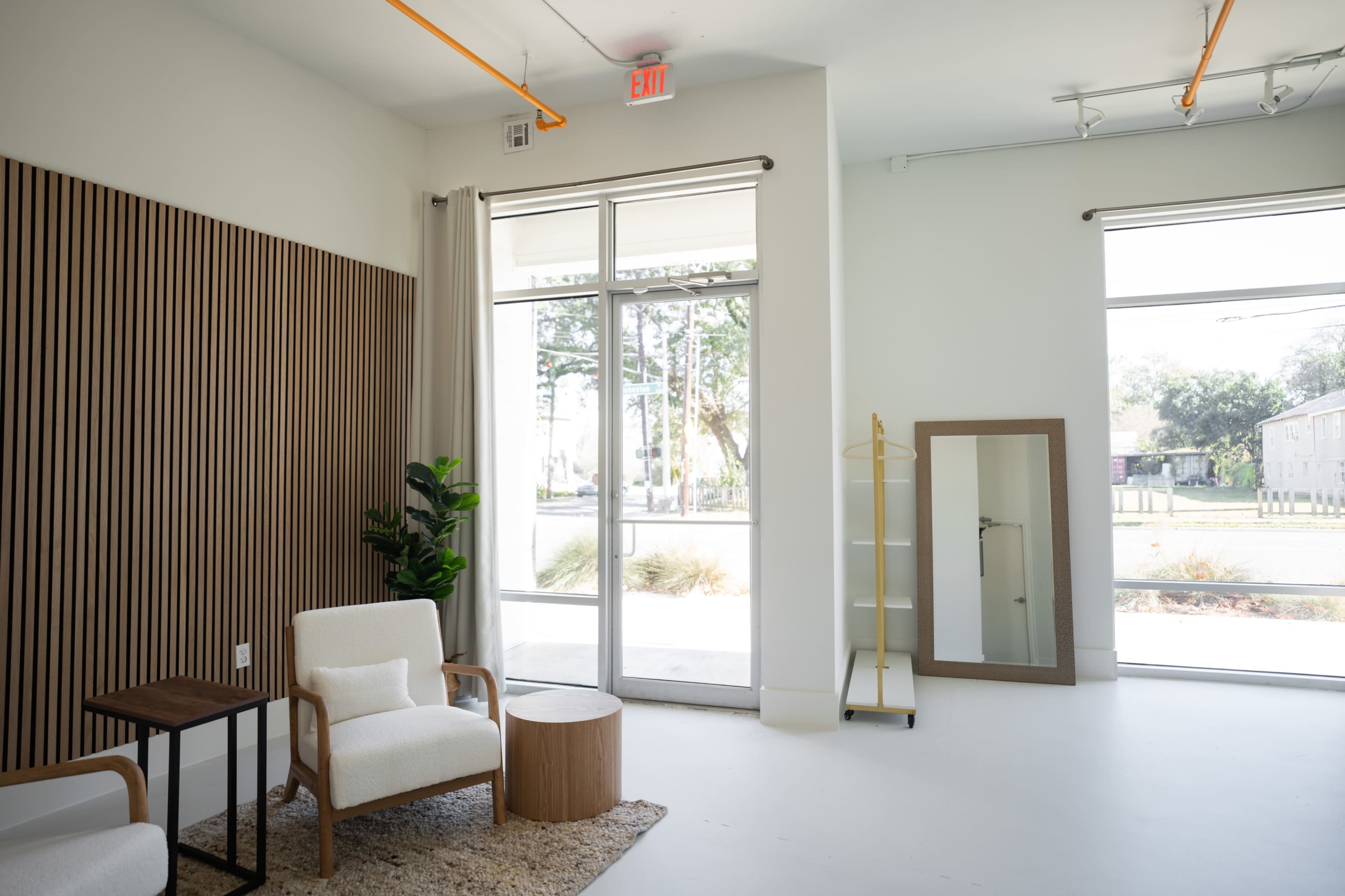 A bright, minimalist interior featuring two armchairs, a small wooden table, a large mirror, and large windows with trees visible outside.