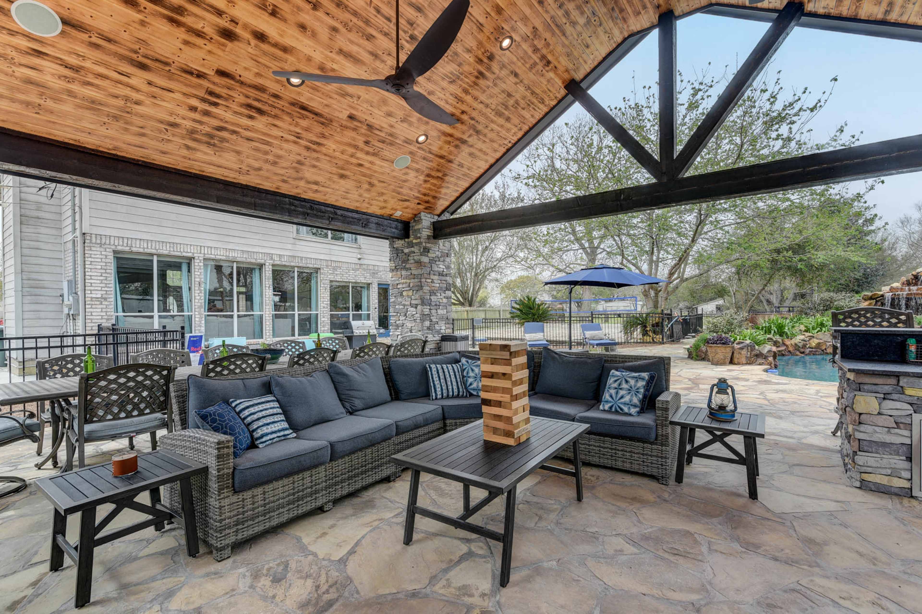 A covered outdoor seating area with a sectional sofa, a coffee table, and a stone barbecue adjacent to a pool area.