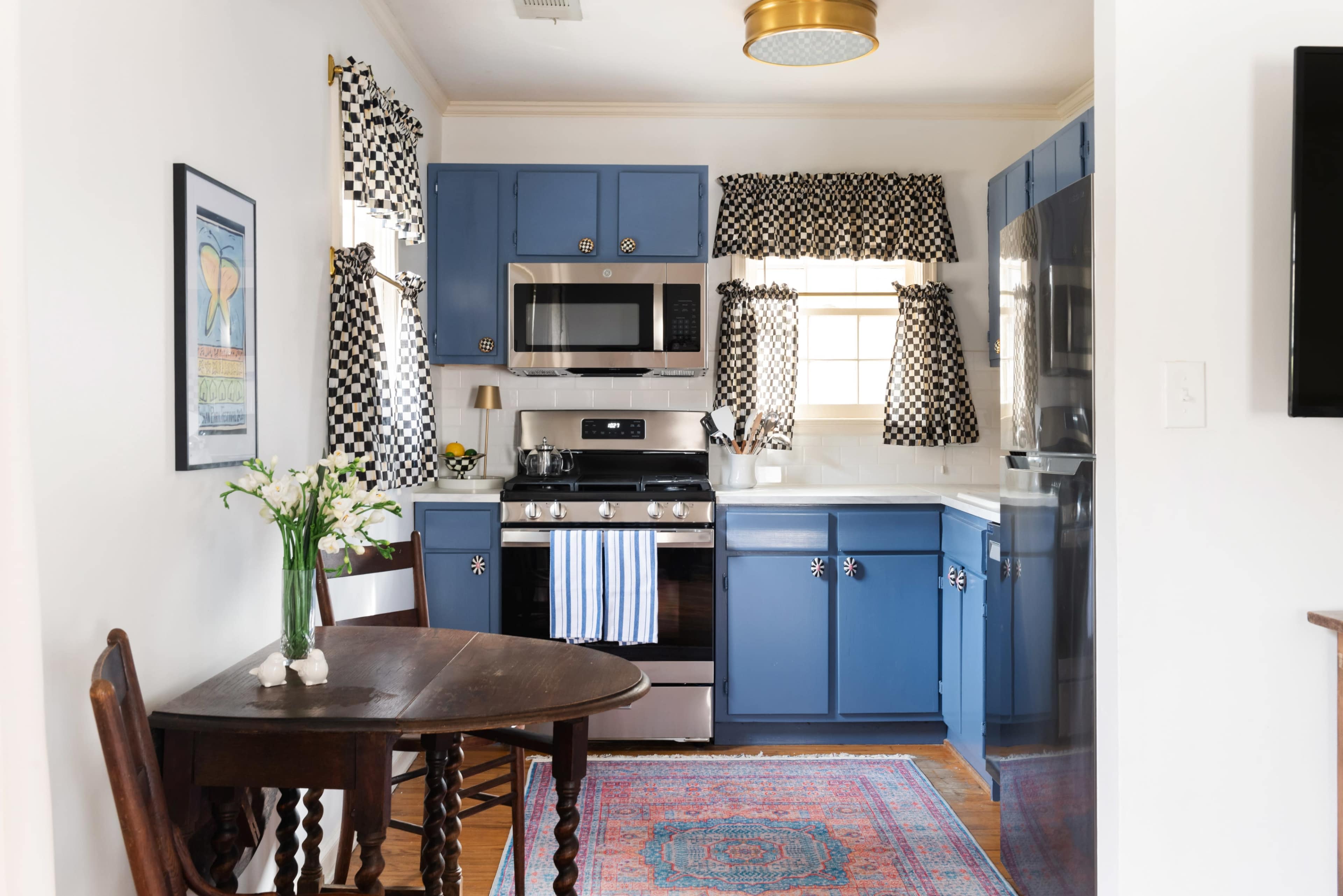 The image shows a kitchen with blue cabinets, a stainless steel stove, and a round wooden table with a vase of flowers, set against a backdrop of checkered curtains.