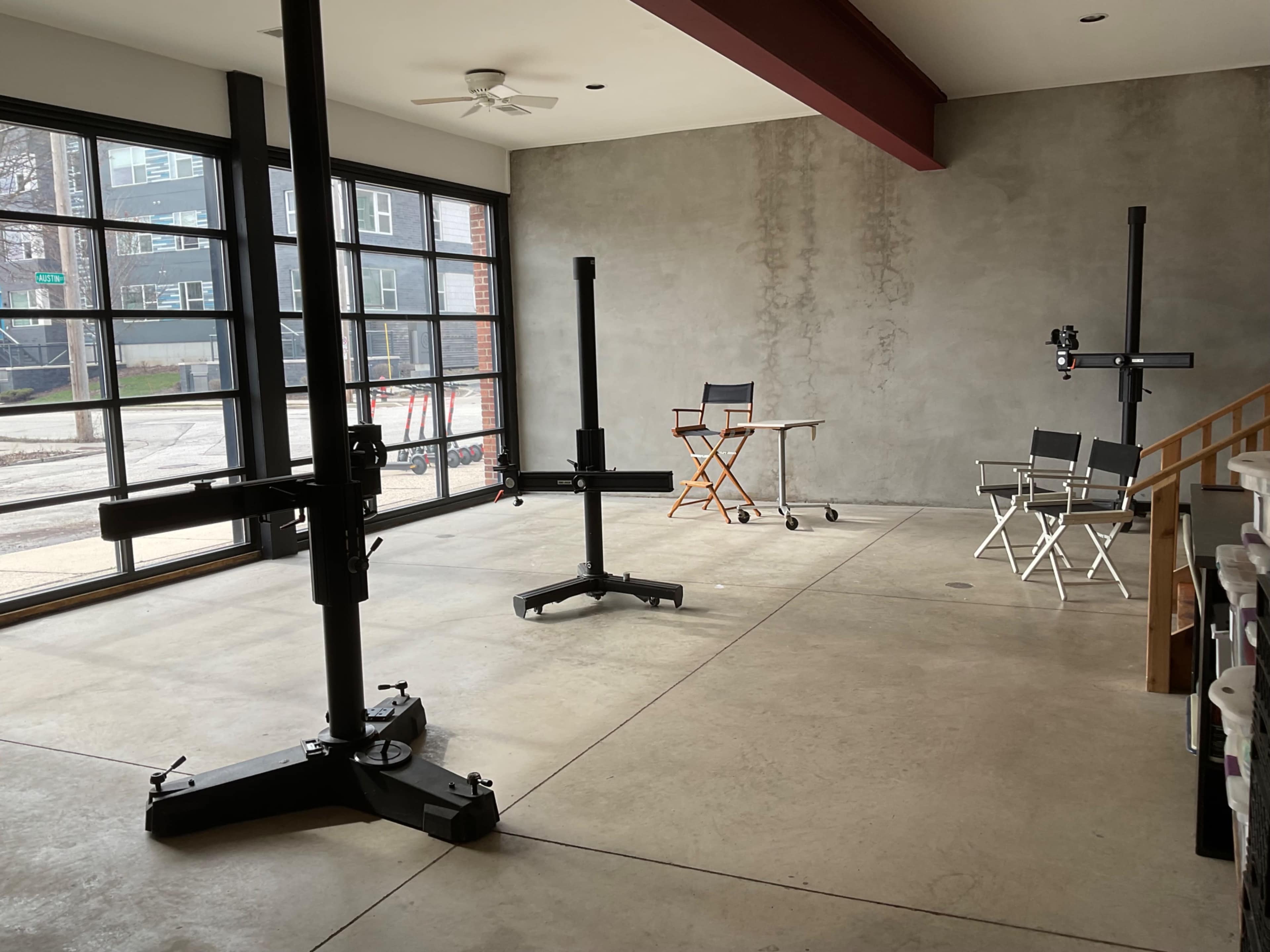 An industrial interior space with concrete floors, large windows, and two film equipment stands, along with a director's chair and a folding chair positioned nearby.