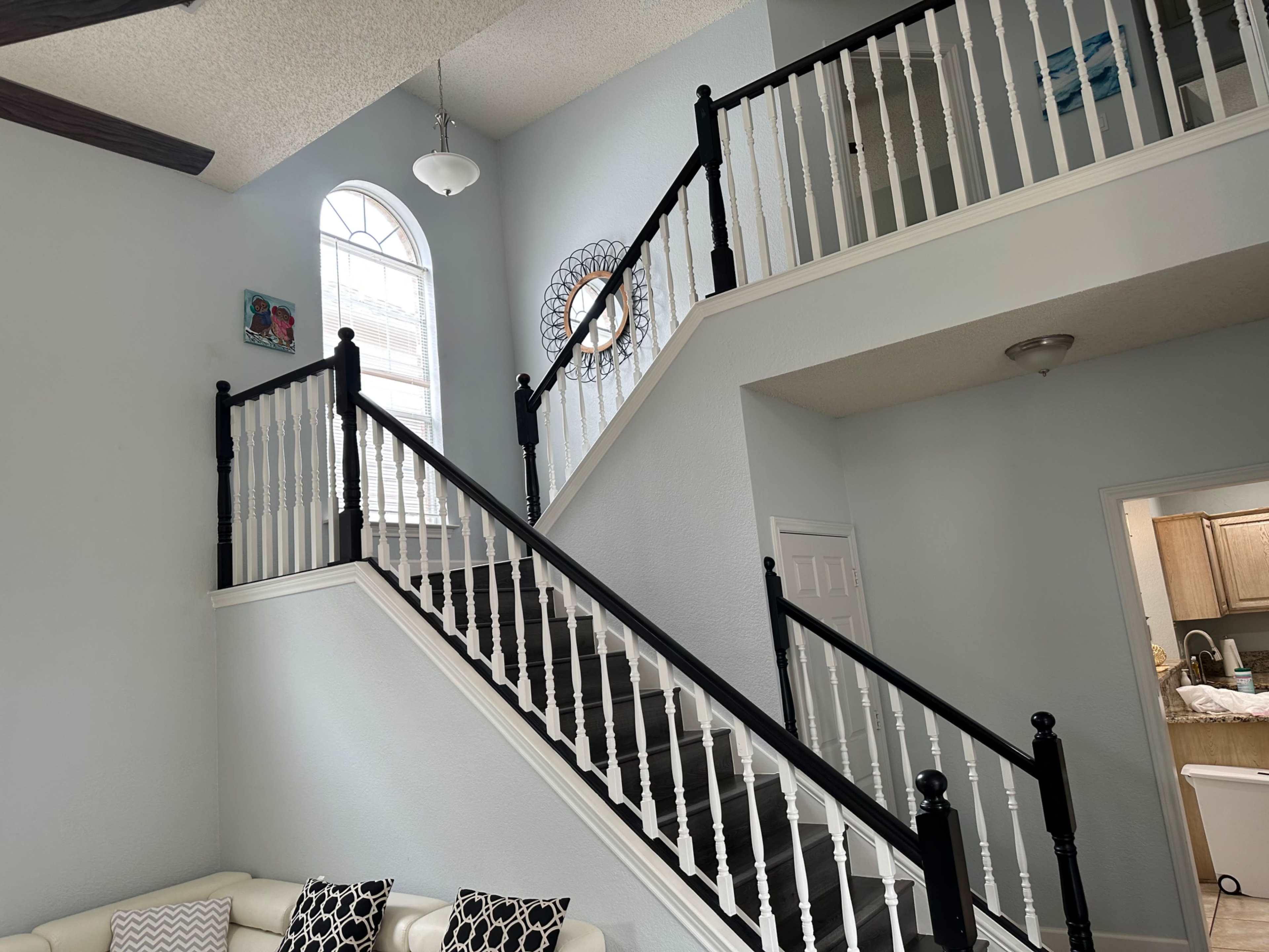 A well-lit living space with a staircase featuring black and white balustrades, leading to a second floor with a circular mirror and a window above.