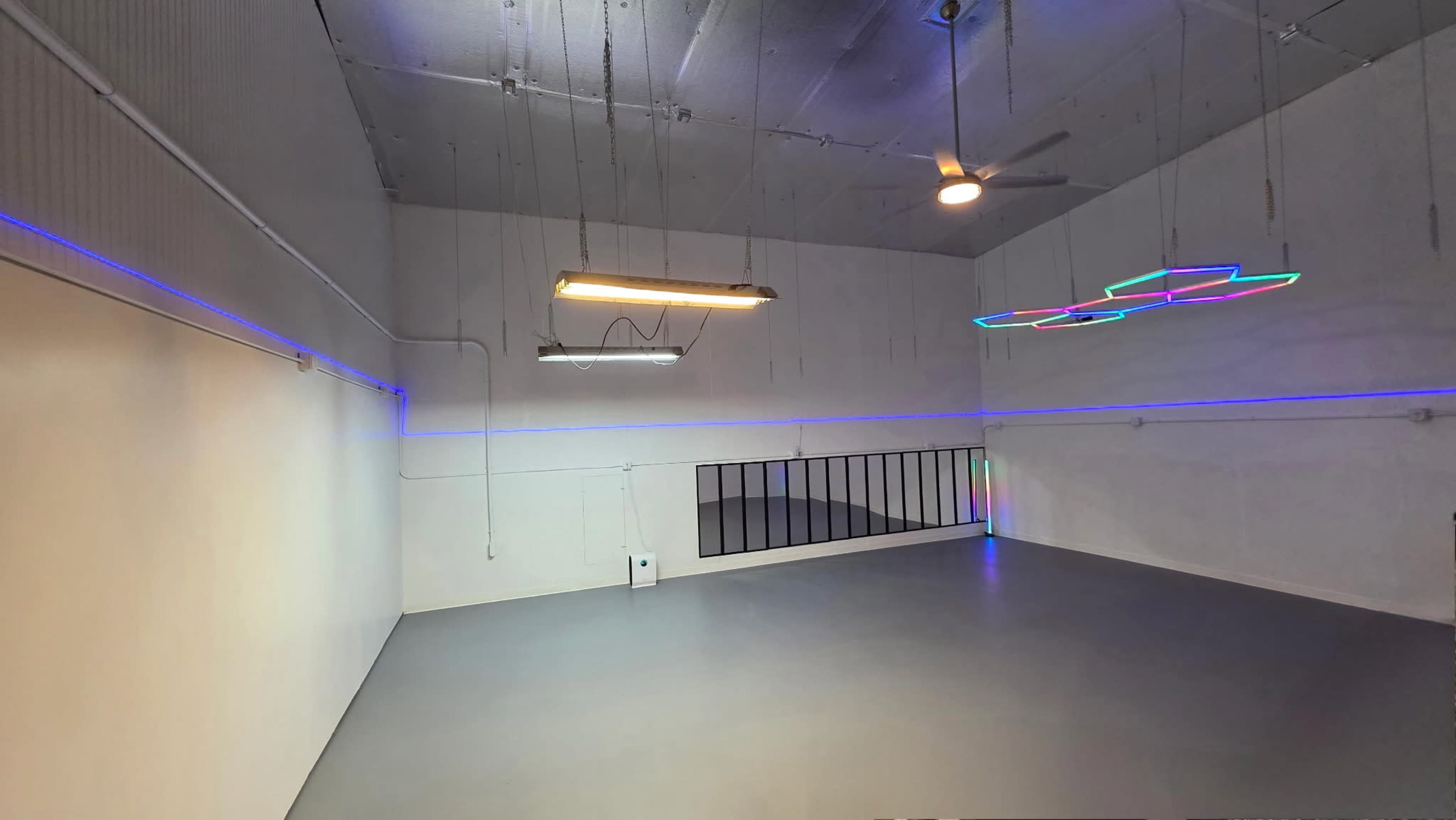 The image shows a spacious, minimalist room with gray flooring, illuminated by colorful LED lights along the walls and a modern light fixture hanging from the ceiling.