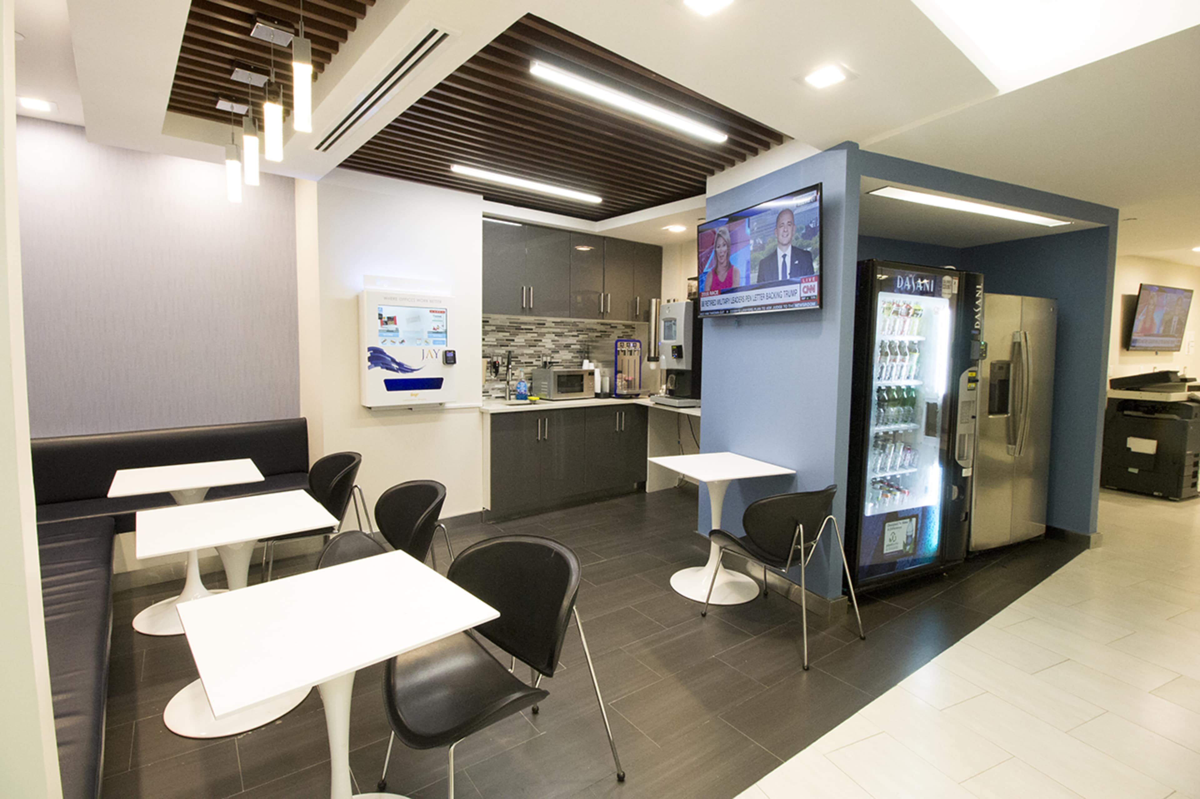 A modern break room with small white tables and black chairs, a kitchen area with cabinets and a television, and a vending machine in the corner.