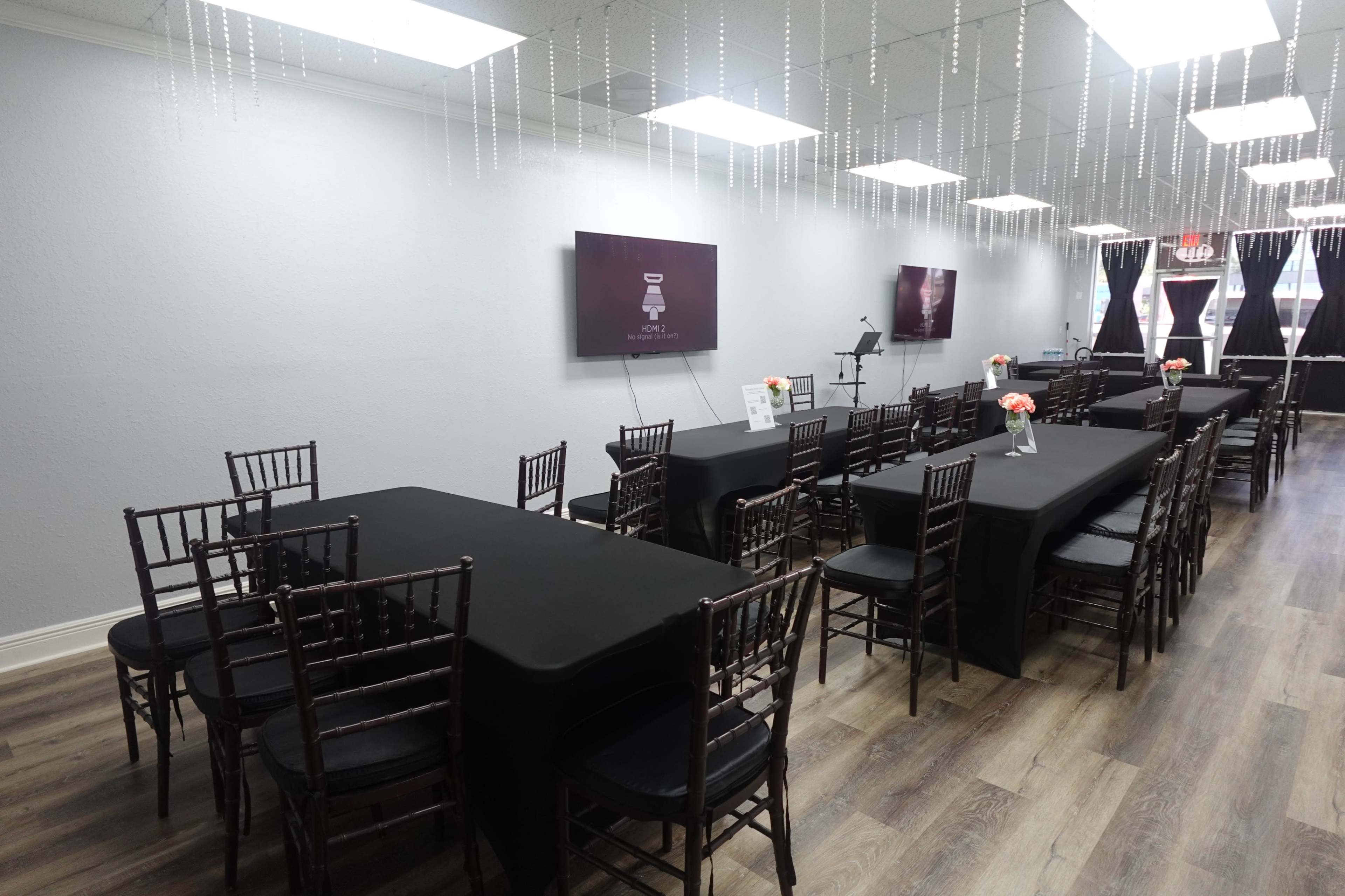 An elegantly arranged event space with long tables covered in black cloth and decorated with small floral centerpieces, flanked by chairs and illuminated by hanging lights.
