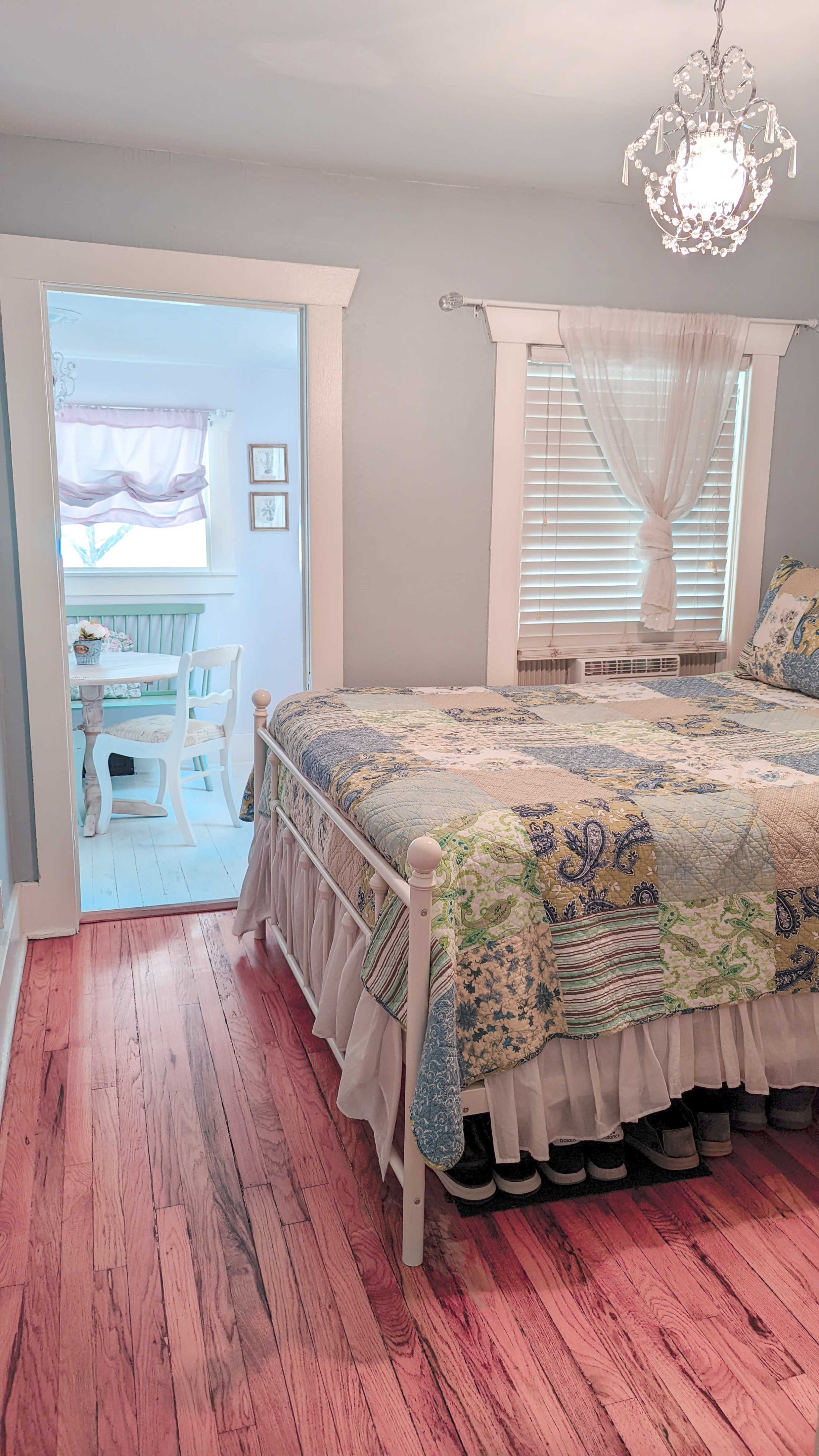 A bedroom with a patterned quilted bedspread, hardwood flooring, and an adjoining room featuring a dining area.