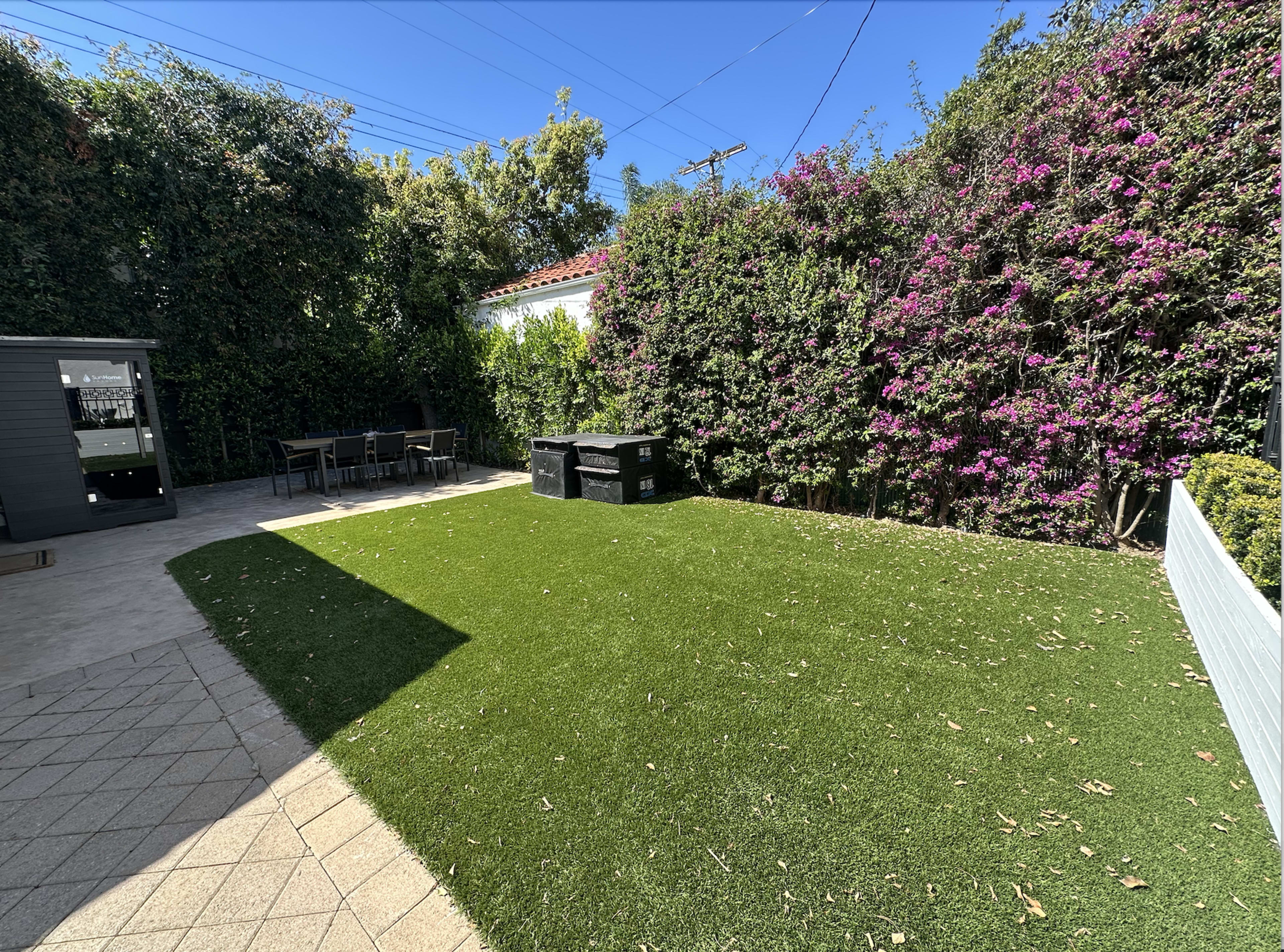 The image shows a landscaped backyard featuring a green lawn, a stone patio, and flowering plants along the perimeter.