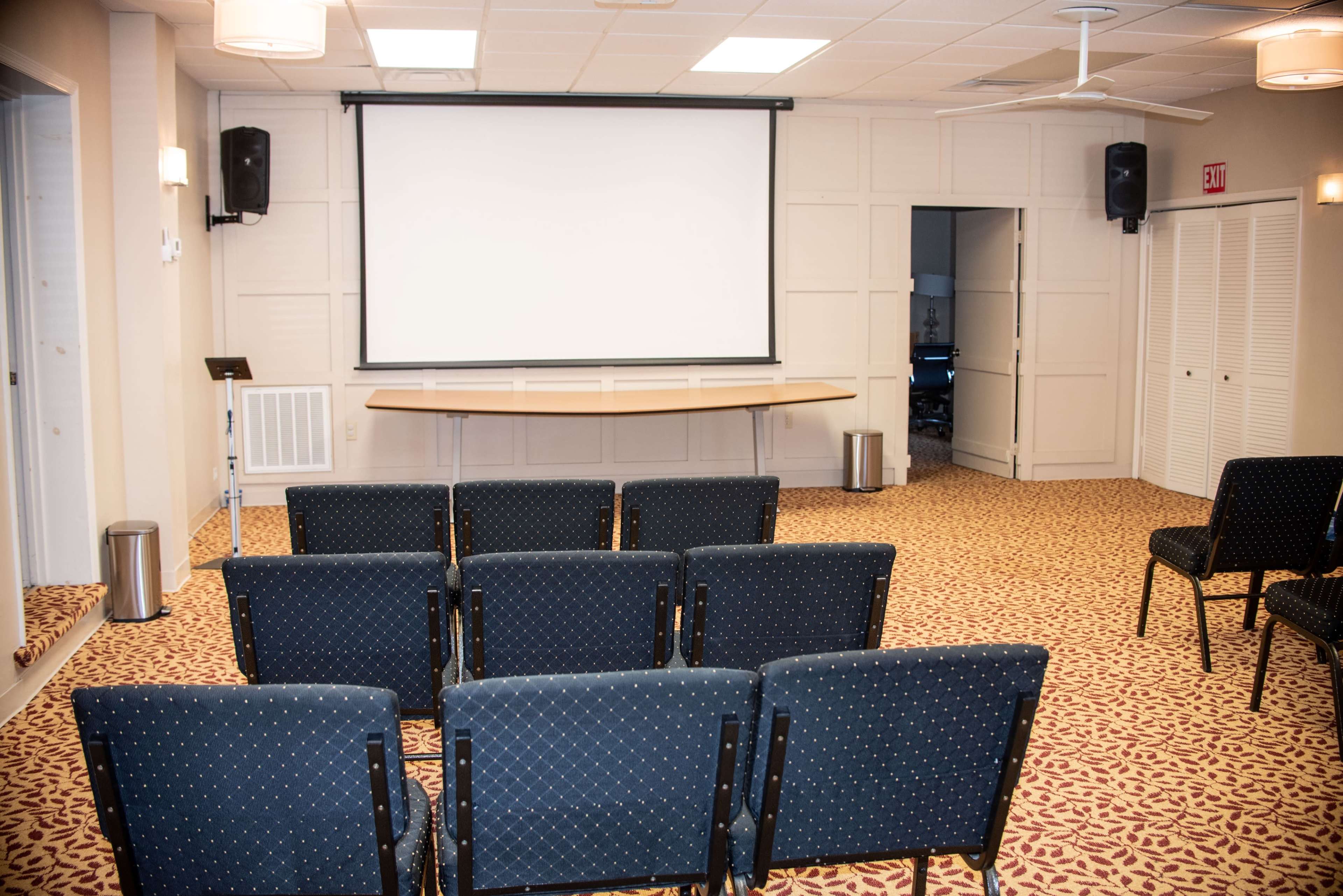 A small conference room with neatly arranged chairs facing a blank projector screen and a door leading to another room.