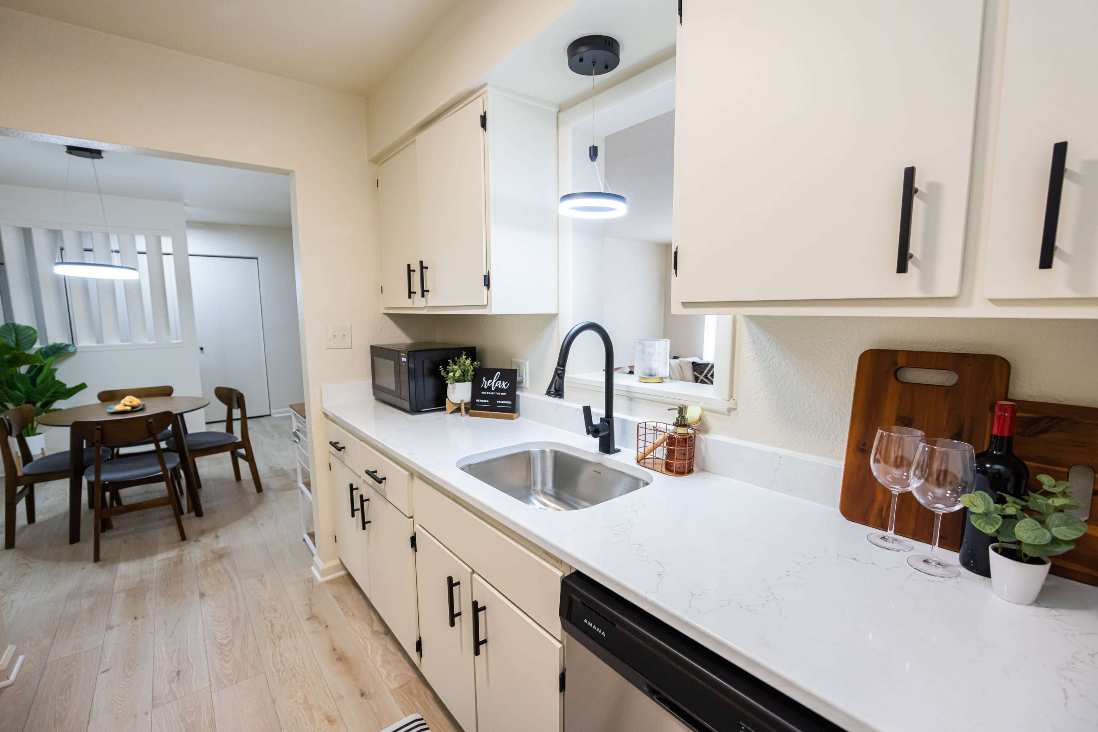 A modern kitchen with white cabinets, a sink, and a countertop, leading to a dining area with a table and chairs visible in the background.