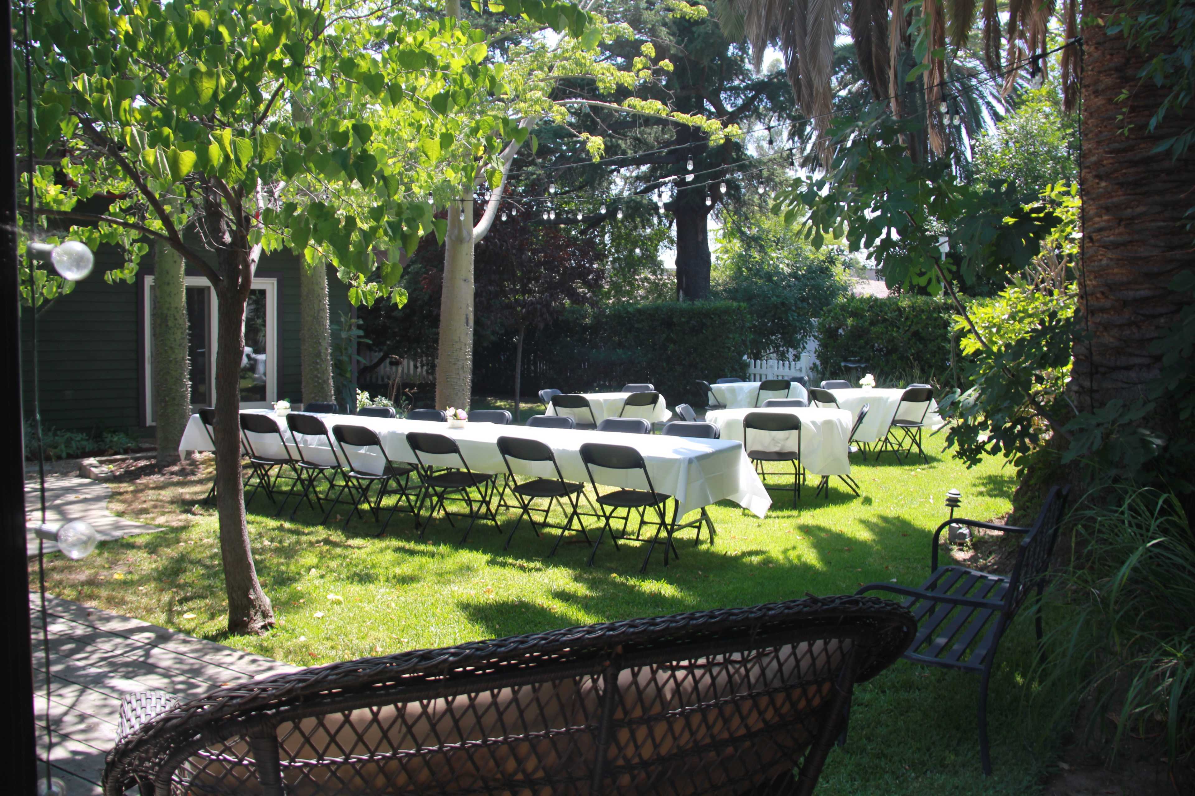 A garden setting features several rows of white-covered tables surrounded by black folding chairs on green grass.