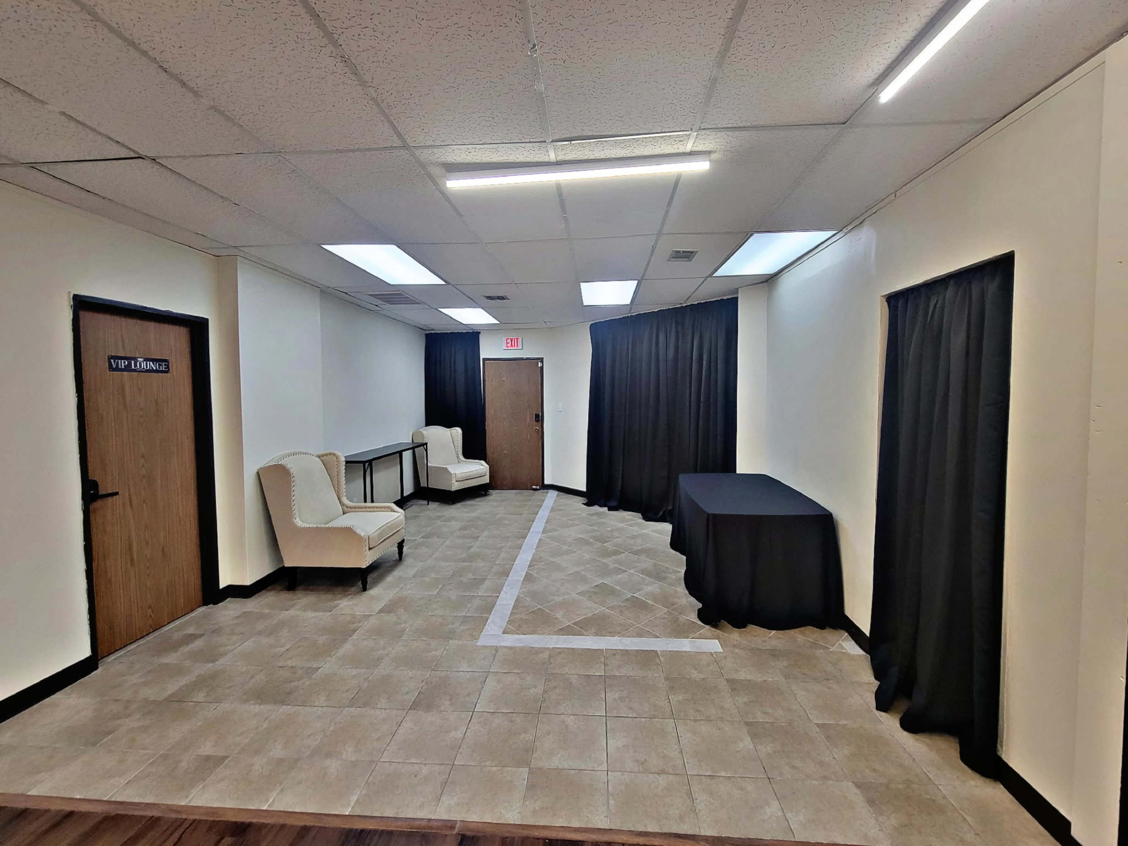 The image shows an empty room with beige tiled flooring, white walls, two light-colored armchairs, a black table, and a backdrop of dark curtains.