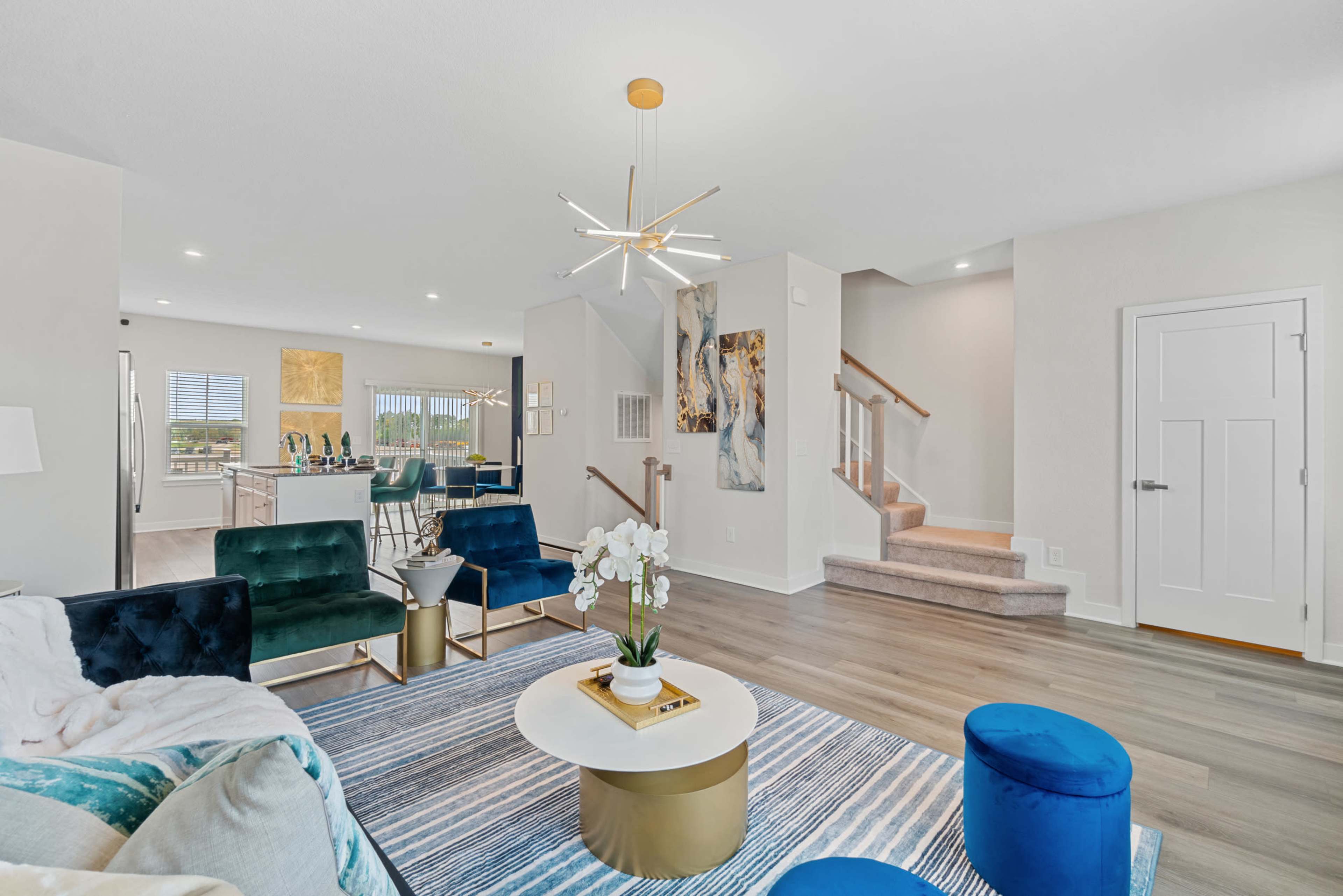 A modern living room with a blue and white area rug, a green and teal seating arrangement, a round coffee table, and a staircase leading to an upper level.