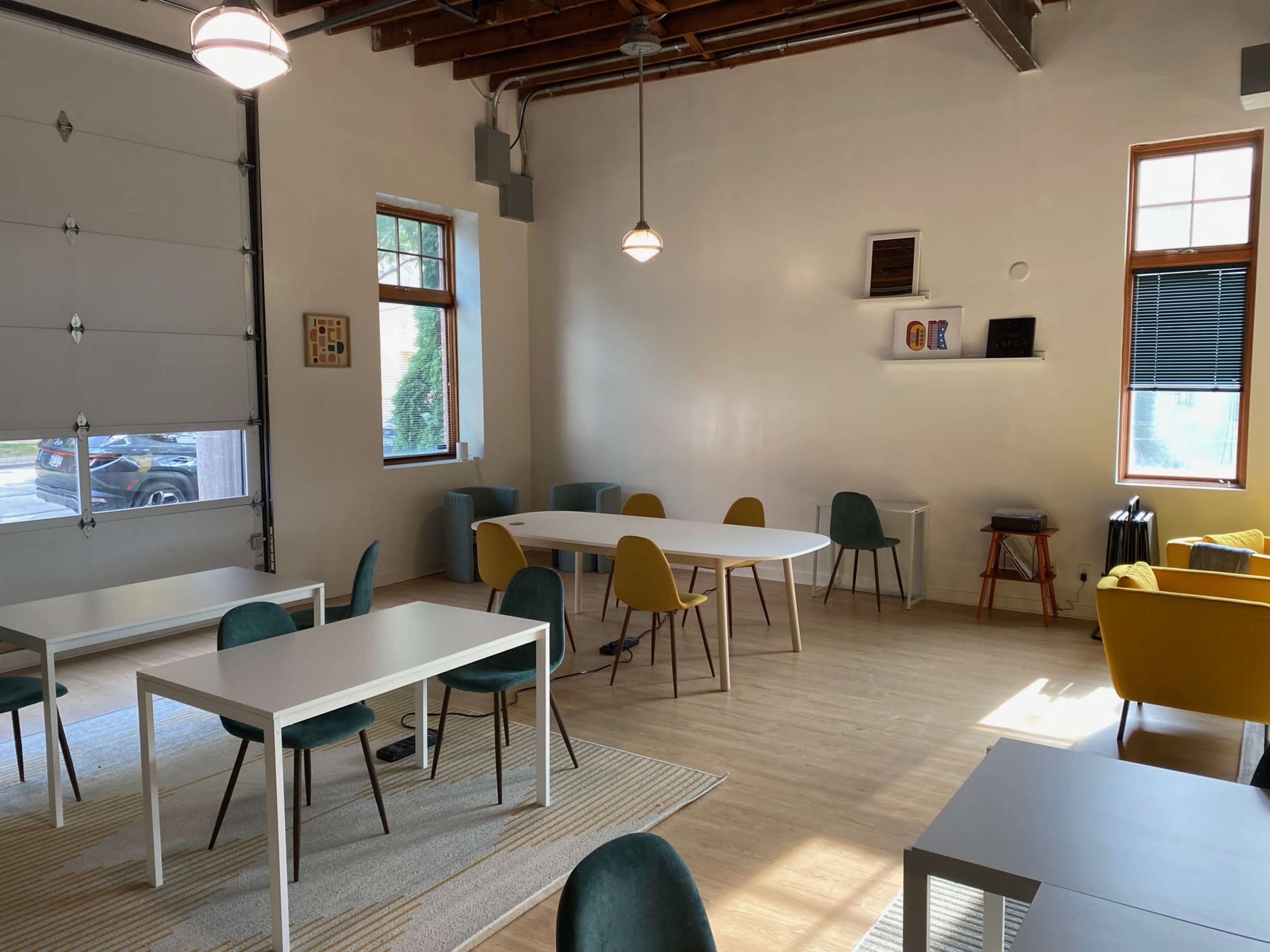 The image shows a modern, open interior space with several tables and chairs, large windows, and a mix of seating areas, including a couch.