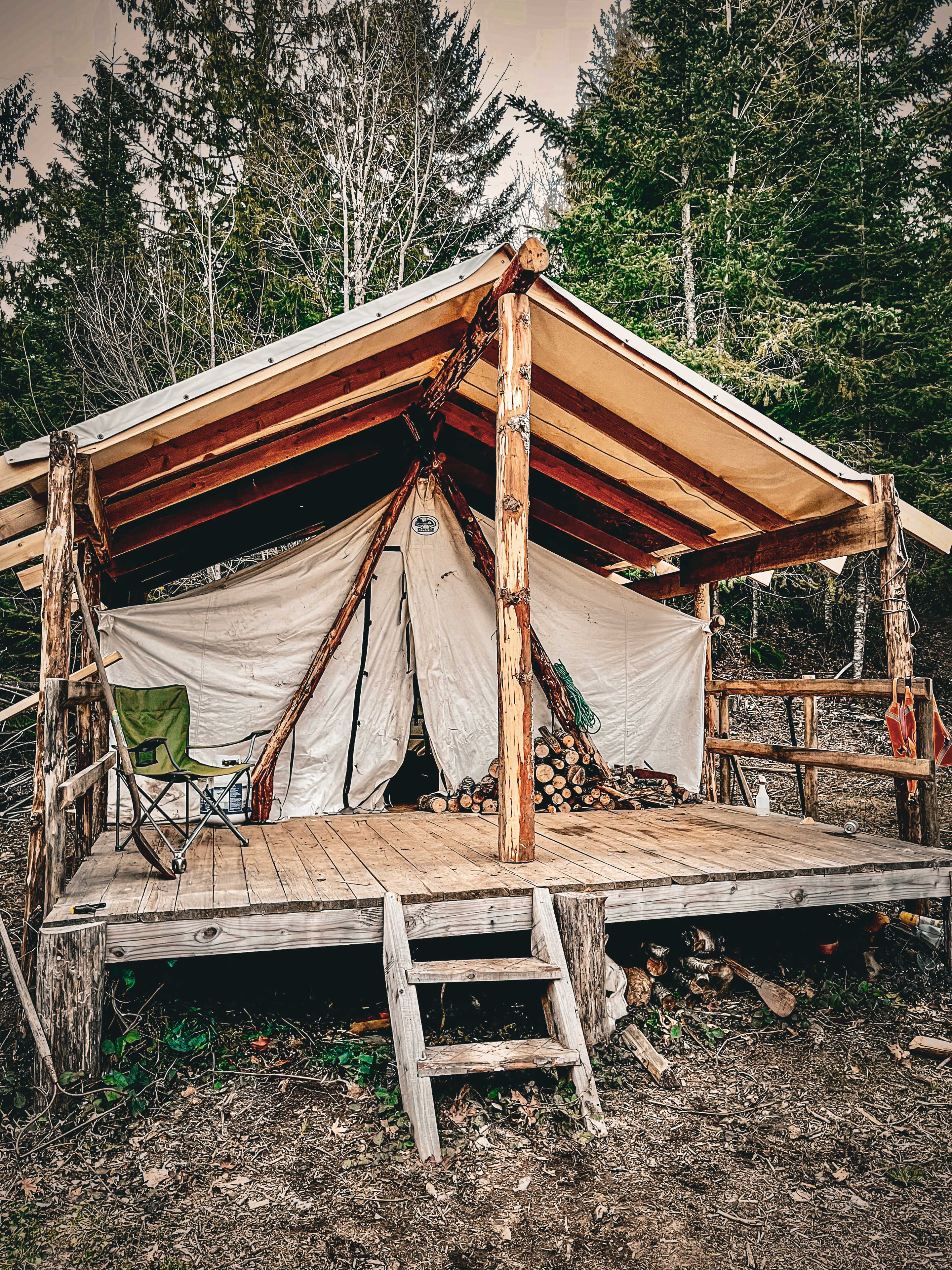 A rustic wooden cabin with a deck is set in a forested area, featuring a sloped roof and canvas walls, along with a folding chair placed outside.