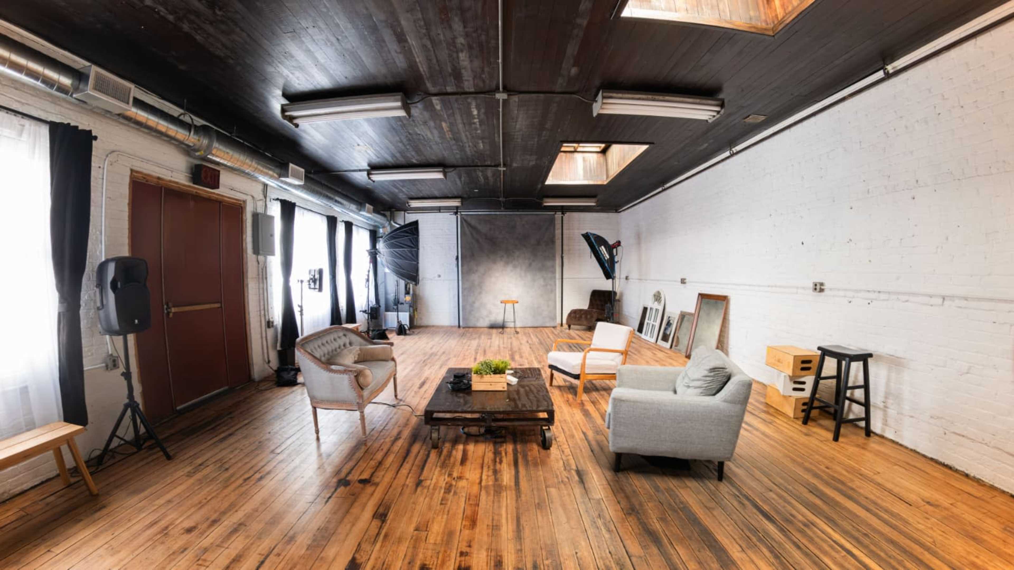 The image shows a spacious room with wooden floors, large windows, and a seating area that includes a gray armchair, a light-colored armchair, and a wooden coffee table.
