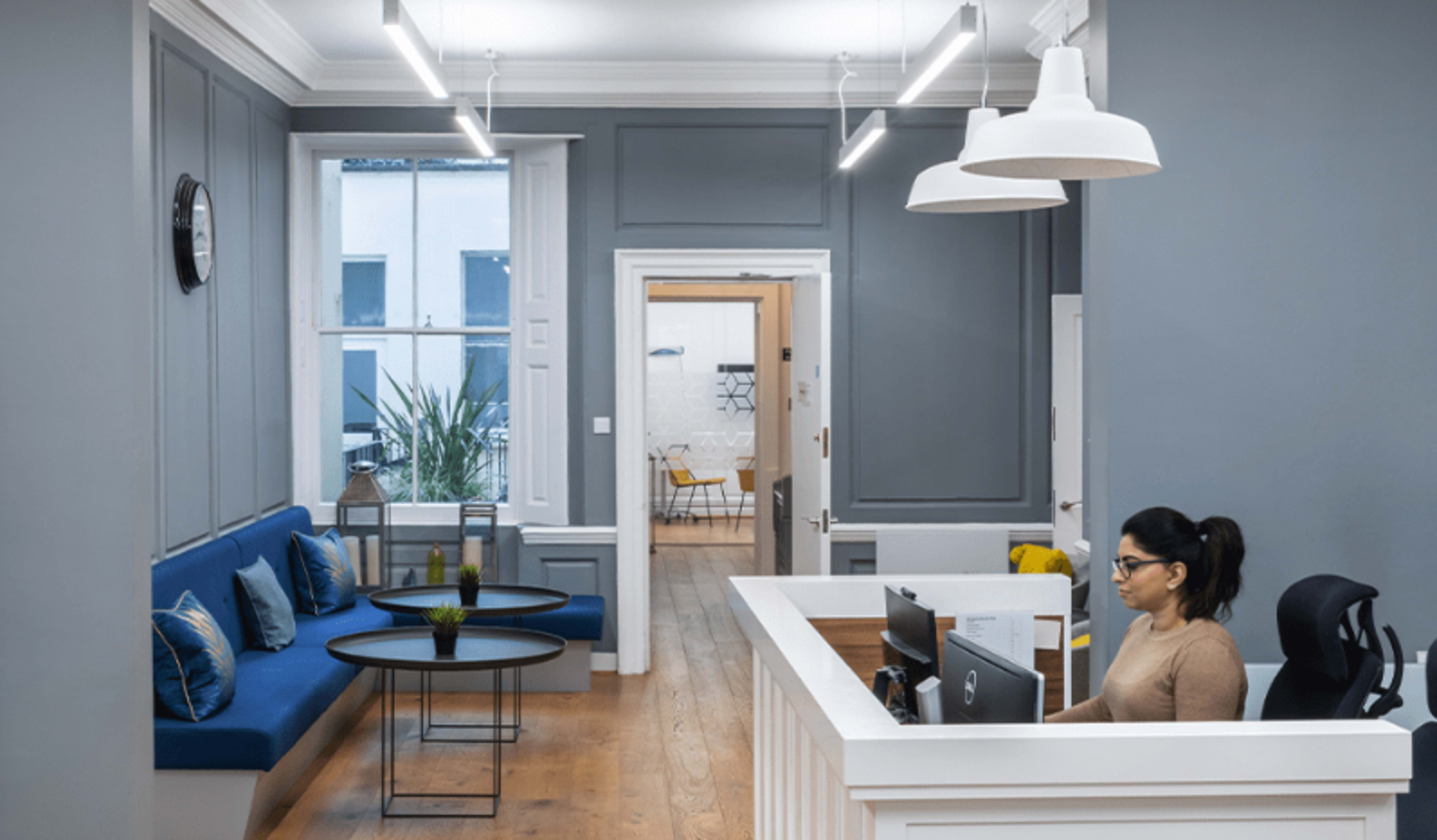 A reception area features a blue sofa, two coffee tables, and a woman working behind a white front desk in a modern office setting.