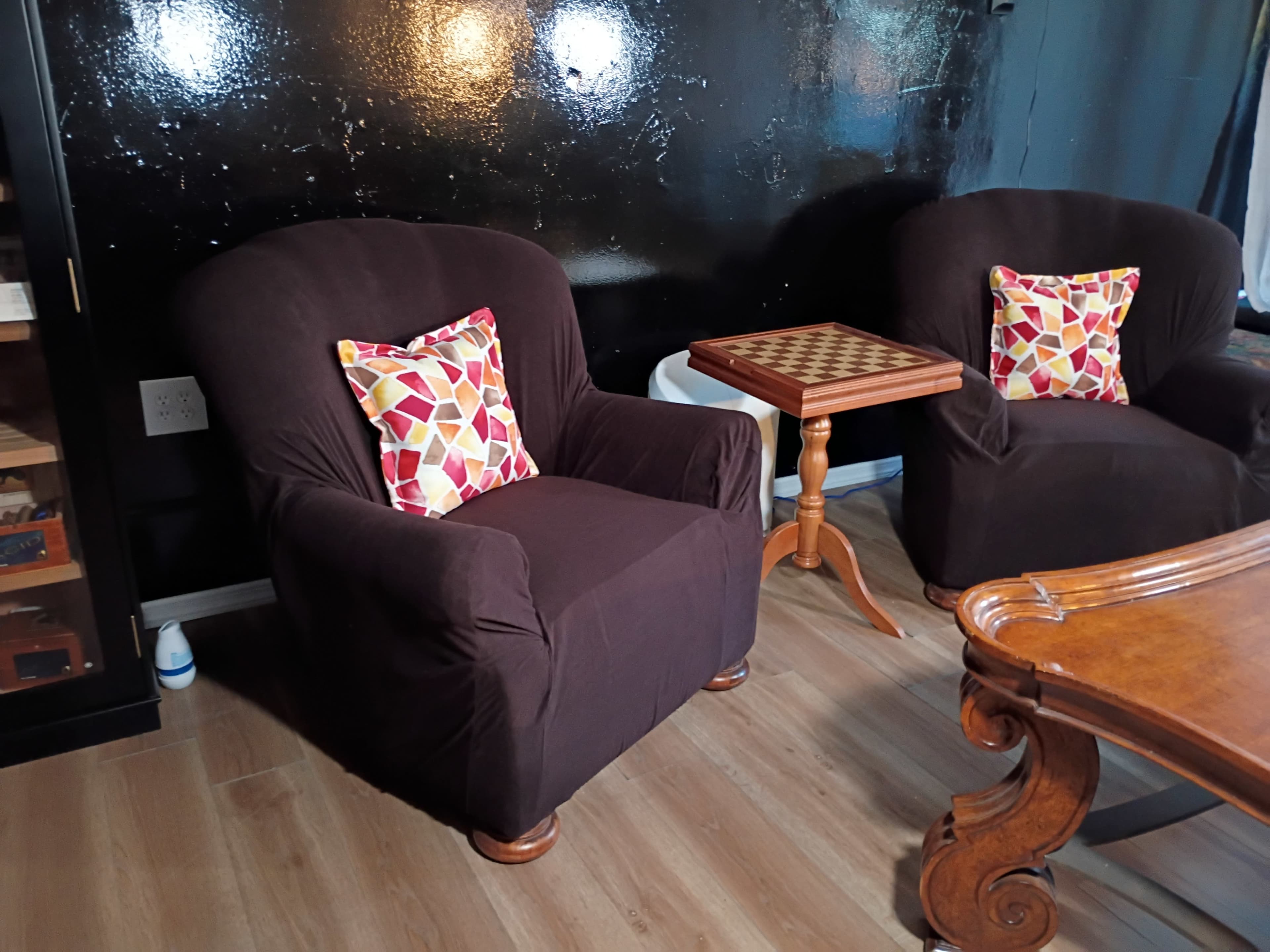 The image shows two dark, upholstered chairs with decorative pillows beside a small wooden side table featuring a checkerboard pattern in a room with a black wall.