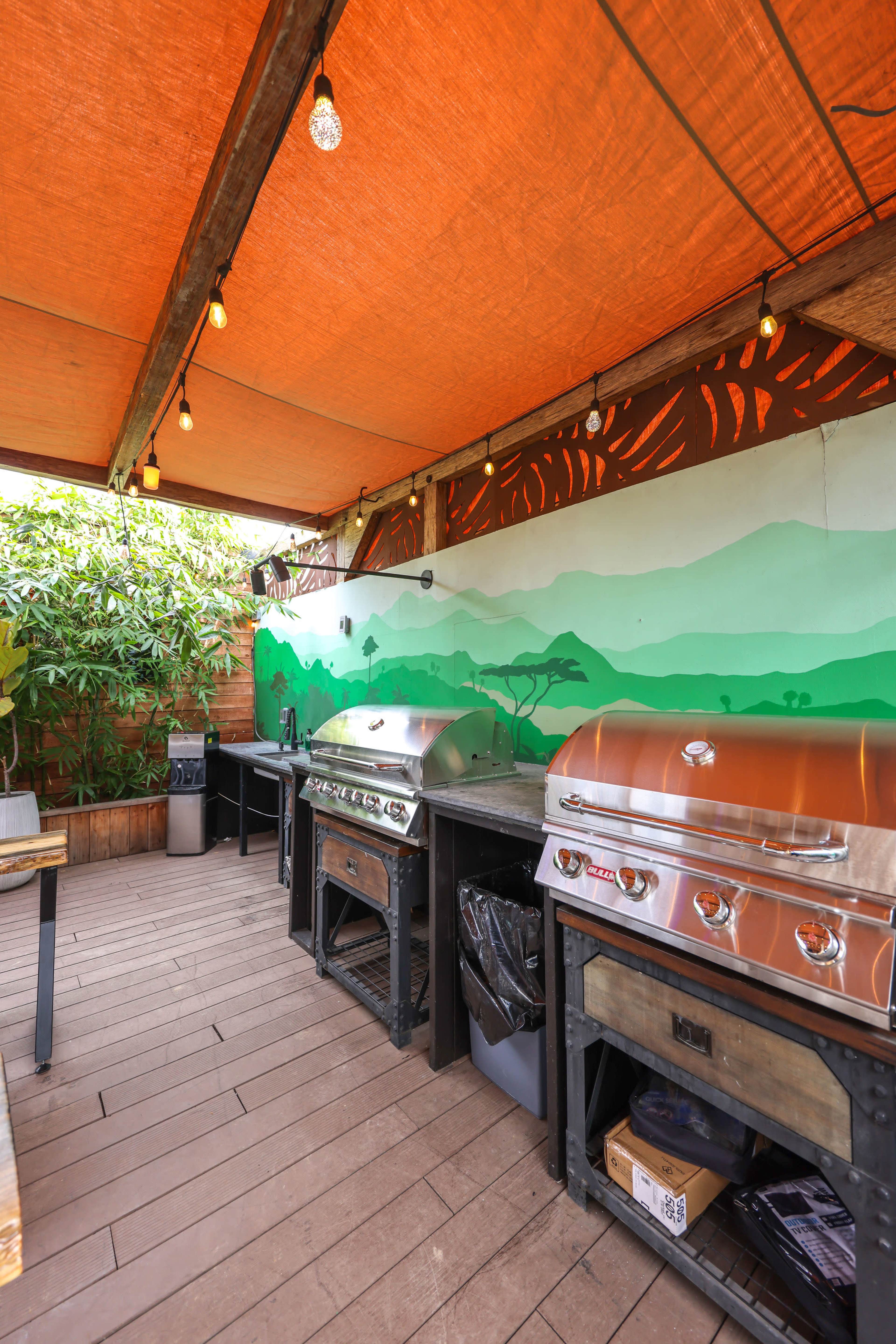 A covered outdoor grilling area with stainless steel grills, wooden countertops, and a mural of green hills on the wall.