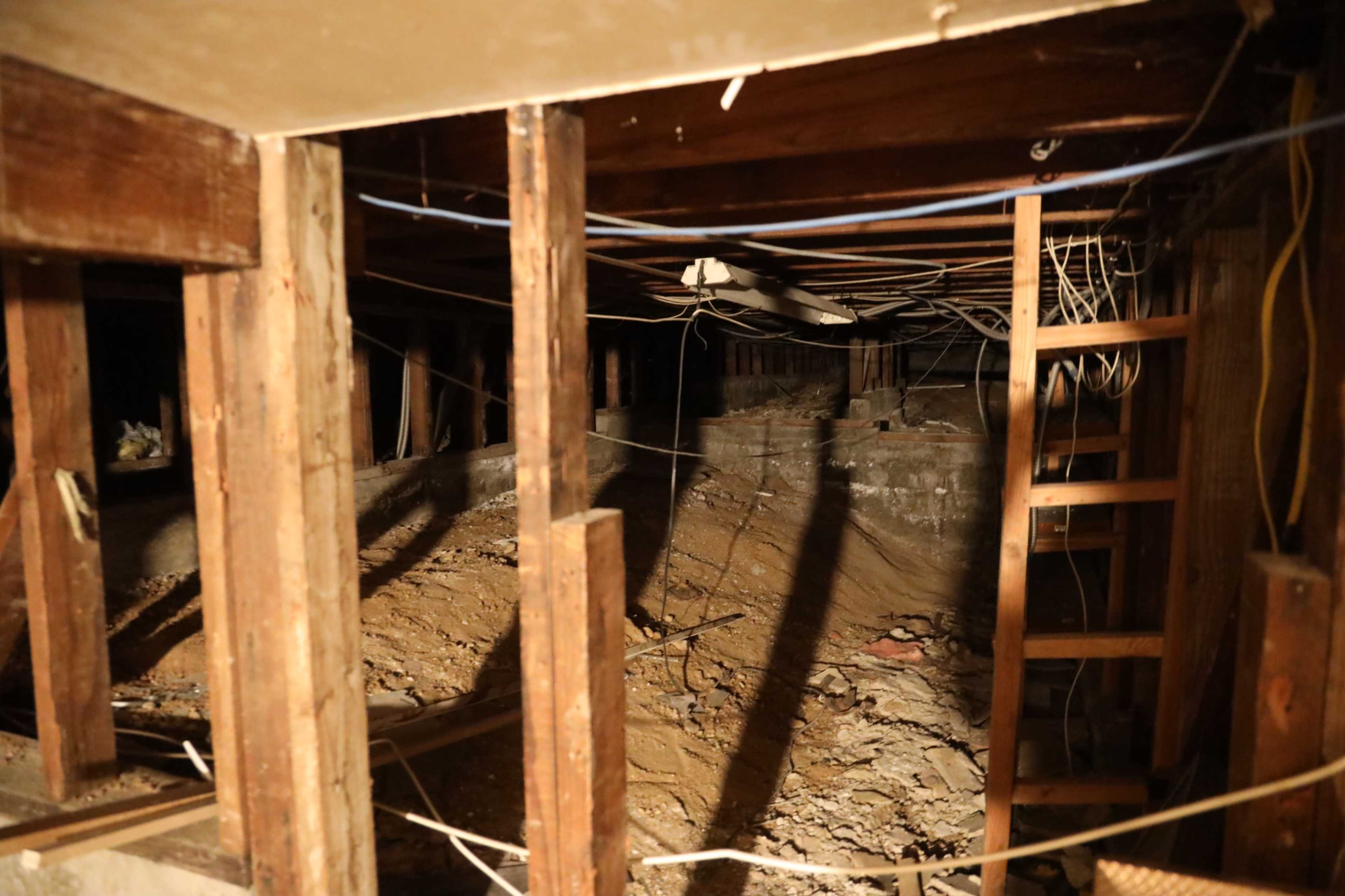 The image shows a dimly lit basement area with exposed wooden beams, scattered debris, and electrical wires running along the ceiling.