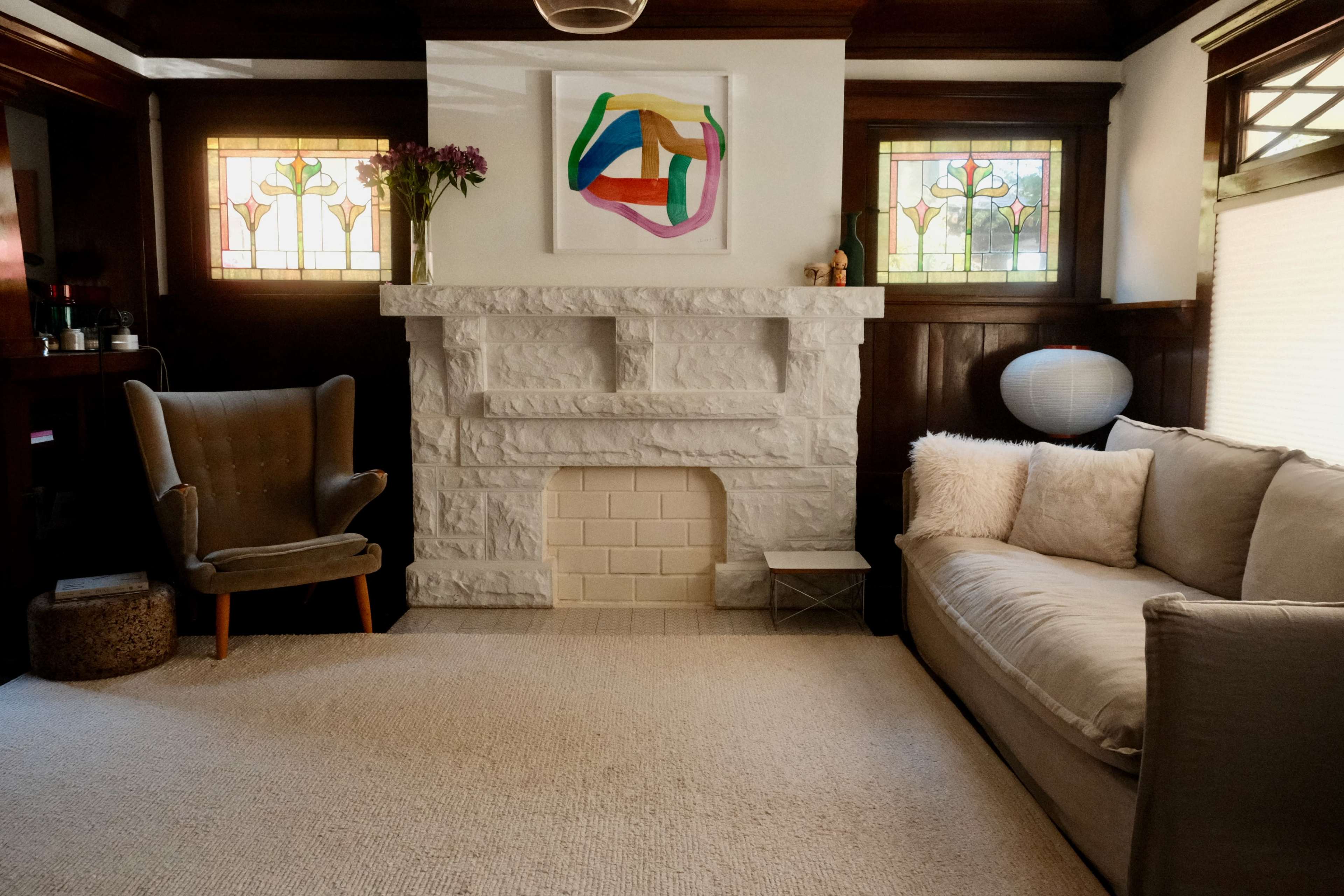 A living room features a light-colored stone fireplace, a gray sofa, a vintage armchair, and stained glass windows.