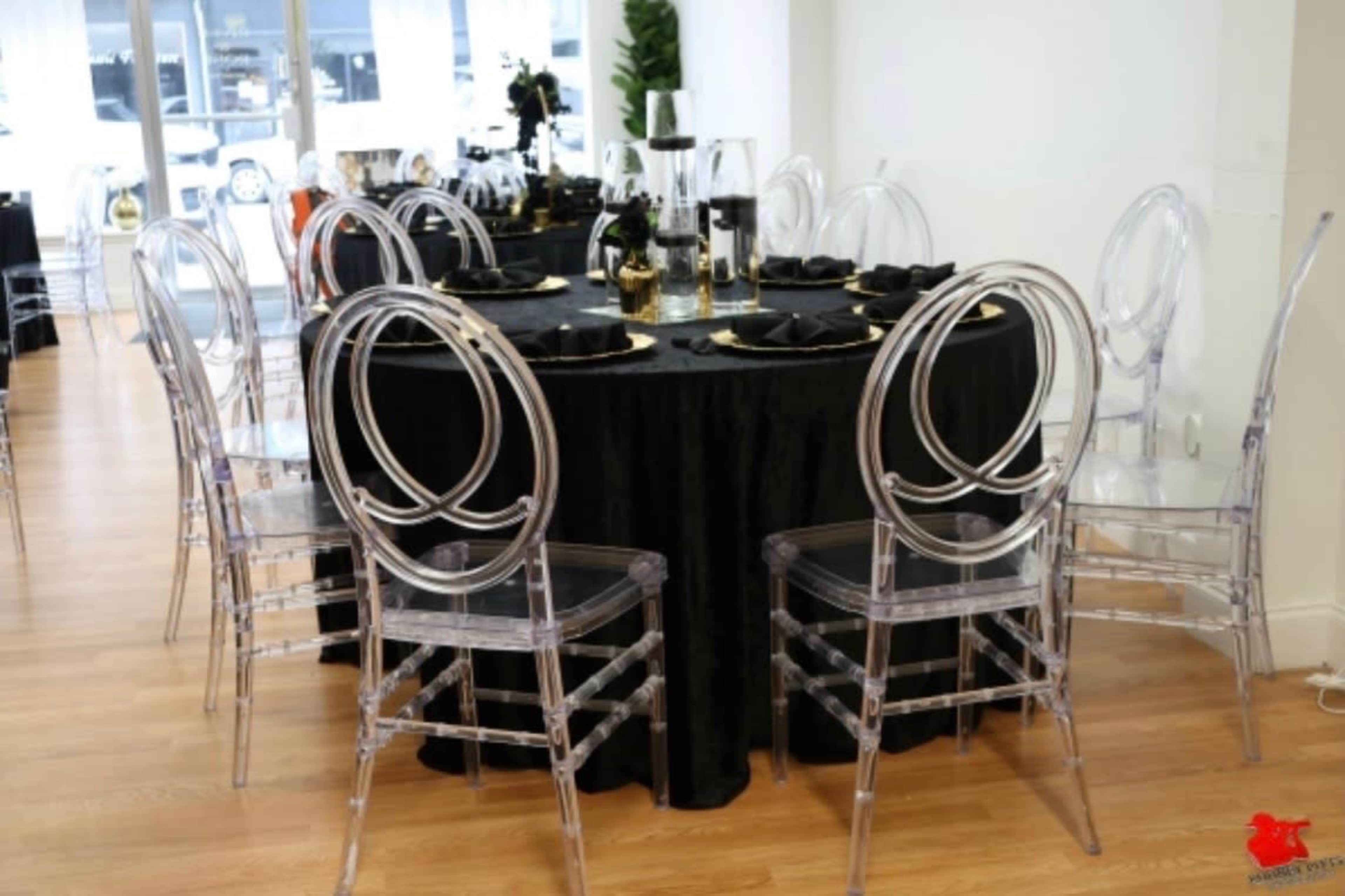 A round table set with black linens and clear acrylic chairs is arranged for dining in a well-lit space.