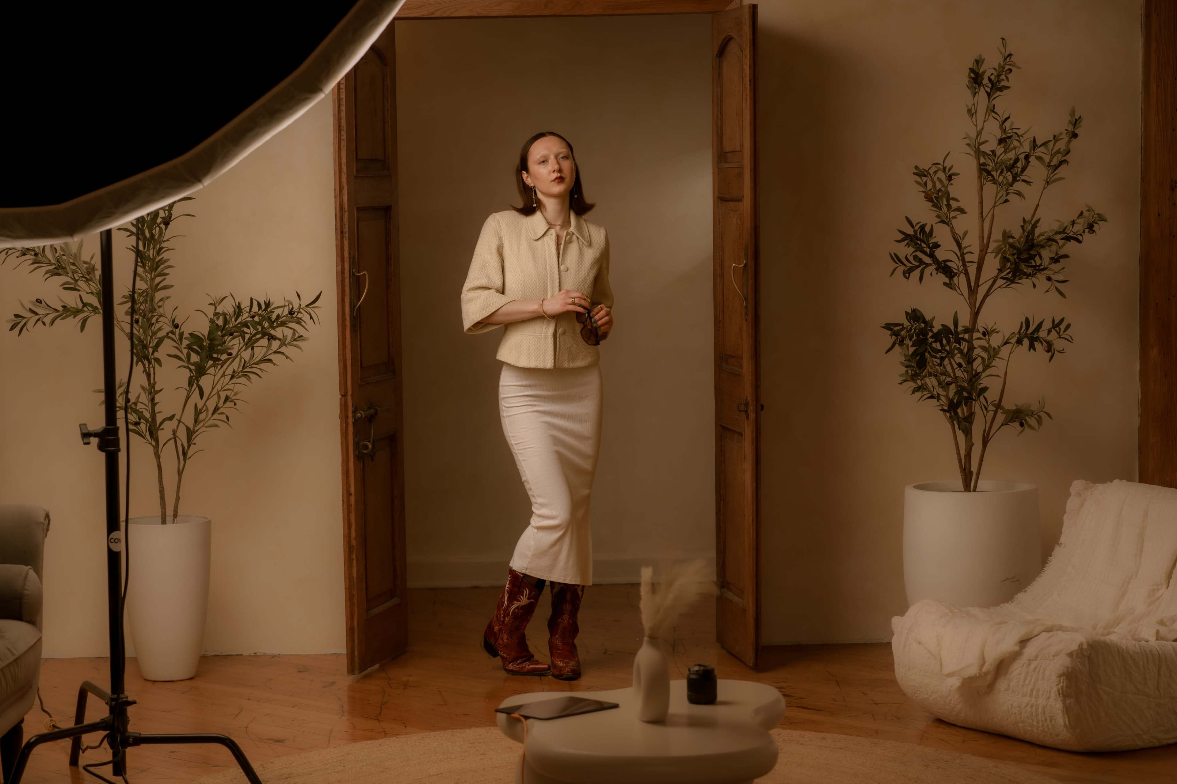 A woman stands in a softly lit room with wooden doors, dressed in a cream-colored top and a white skirt, surrounded by potted plants and minimalistic furniture.