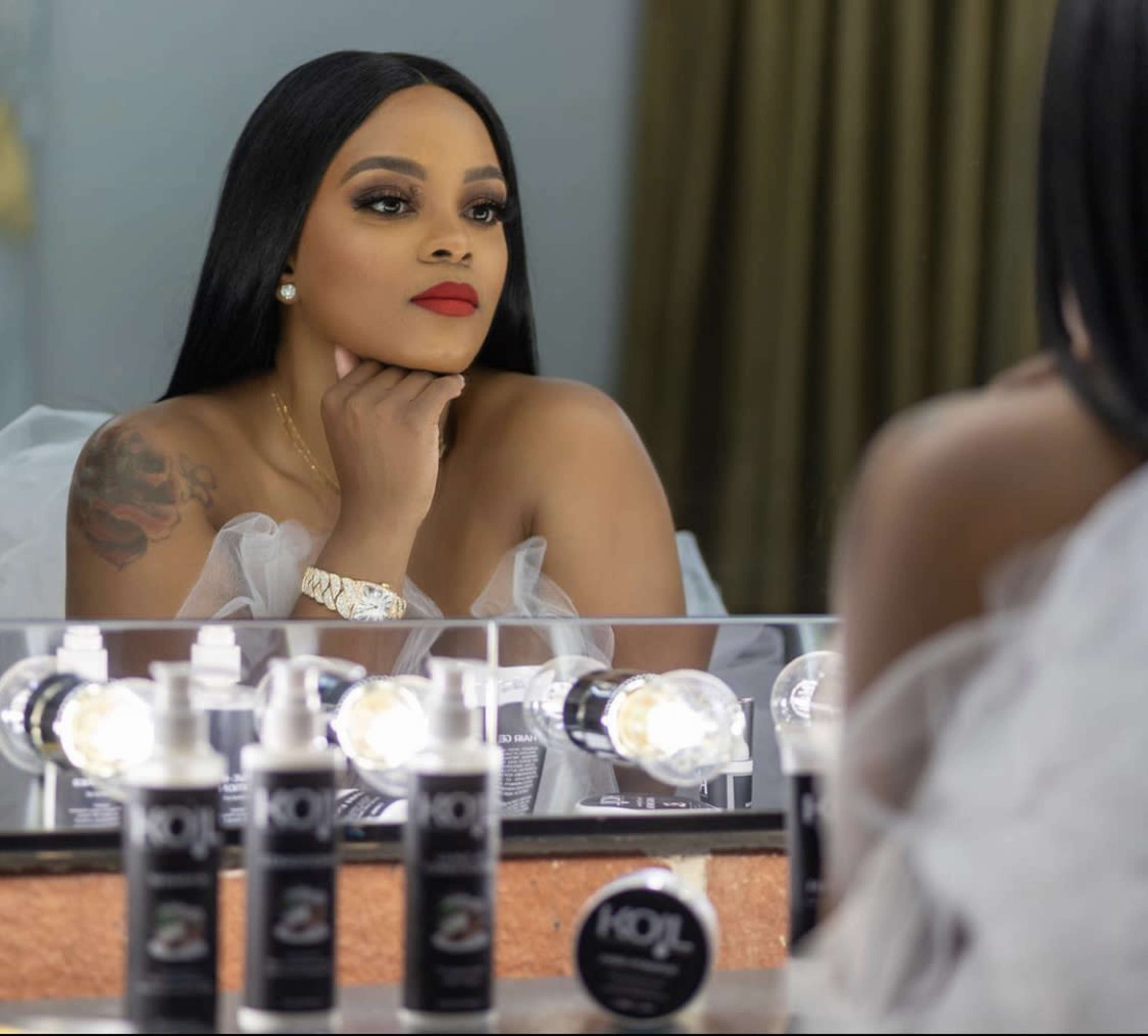 A woman with long black hair and red lipstick gazes at her reflection in a mirror, surrounded by makeup products and illuminated by bright lights.