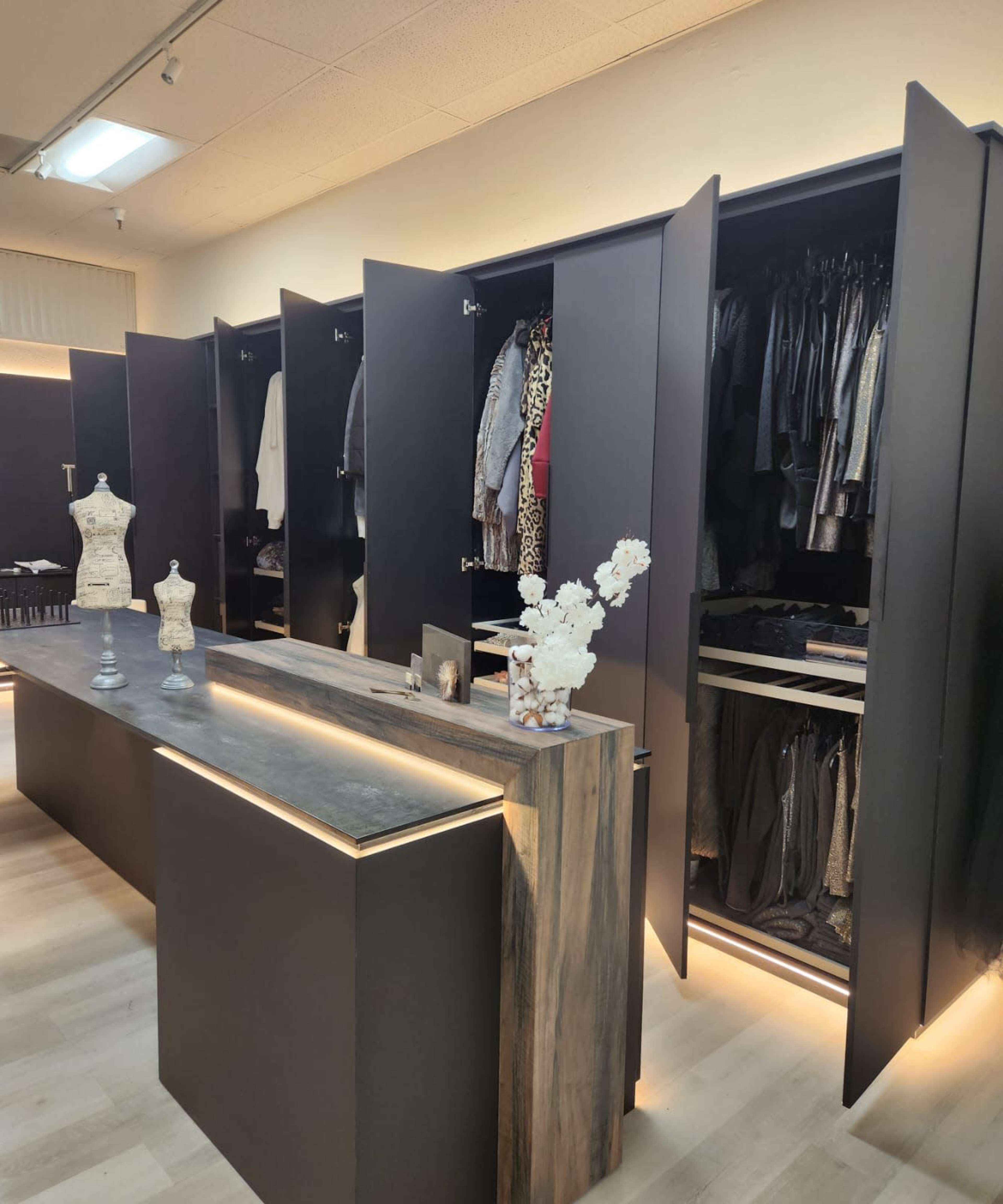 The image shows a modern dressing room with black cabinetry featuring open doors revealing neatly arranged clothes, alongside a wooden counter with decorative items.
