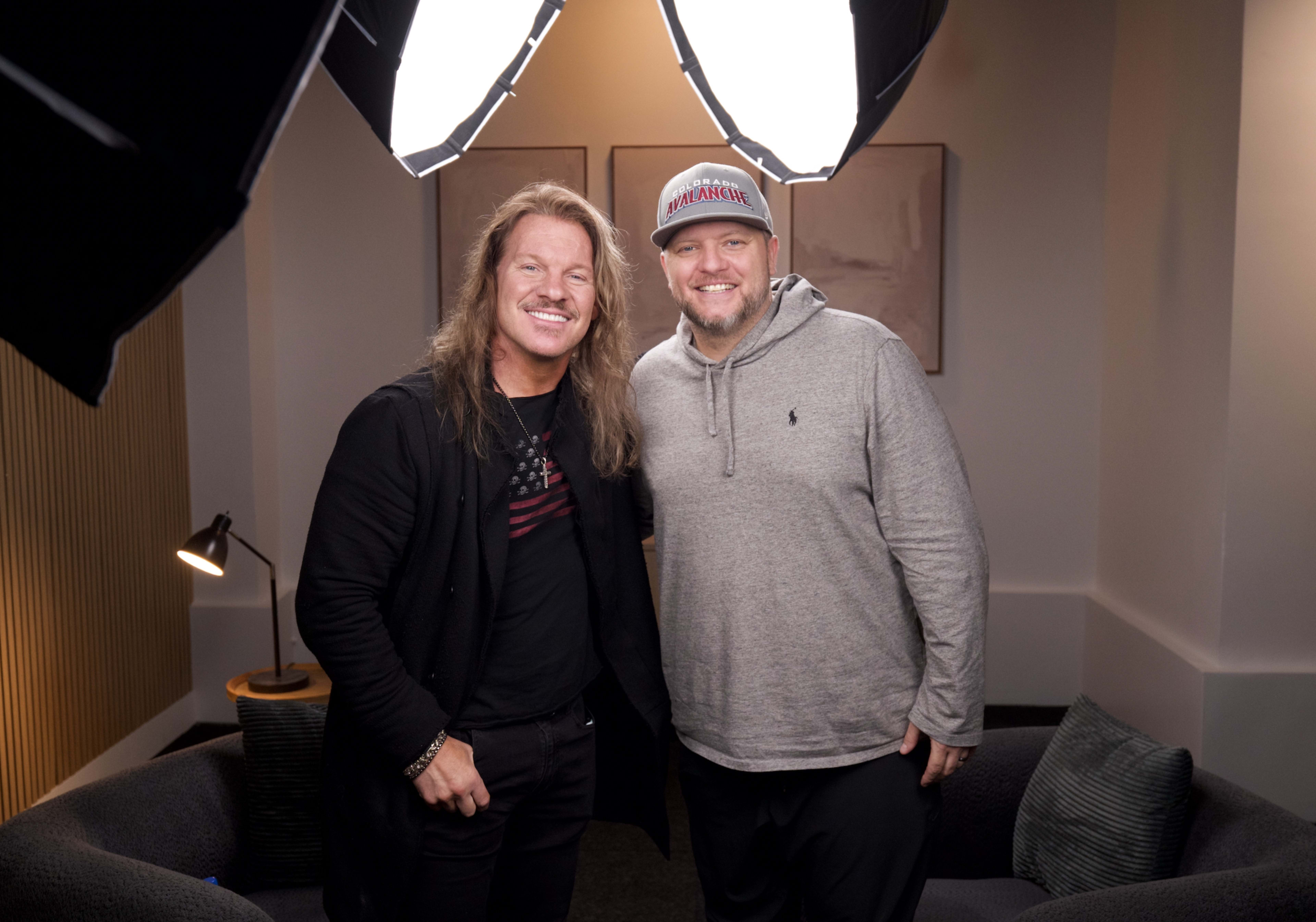 Two men stand together in a cozy setting, smiling at the camera, with studio lighting overhead.