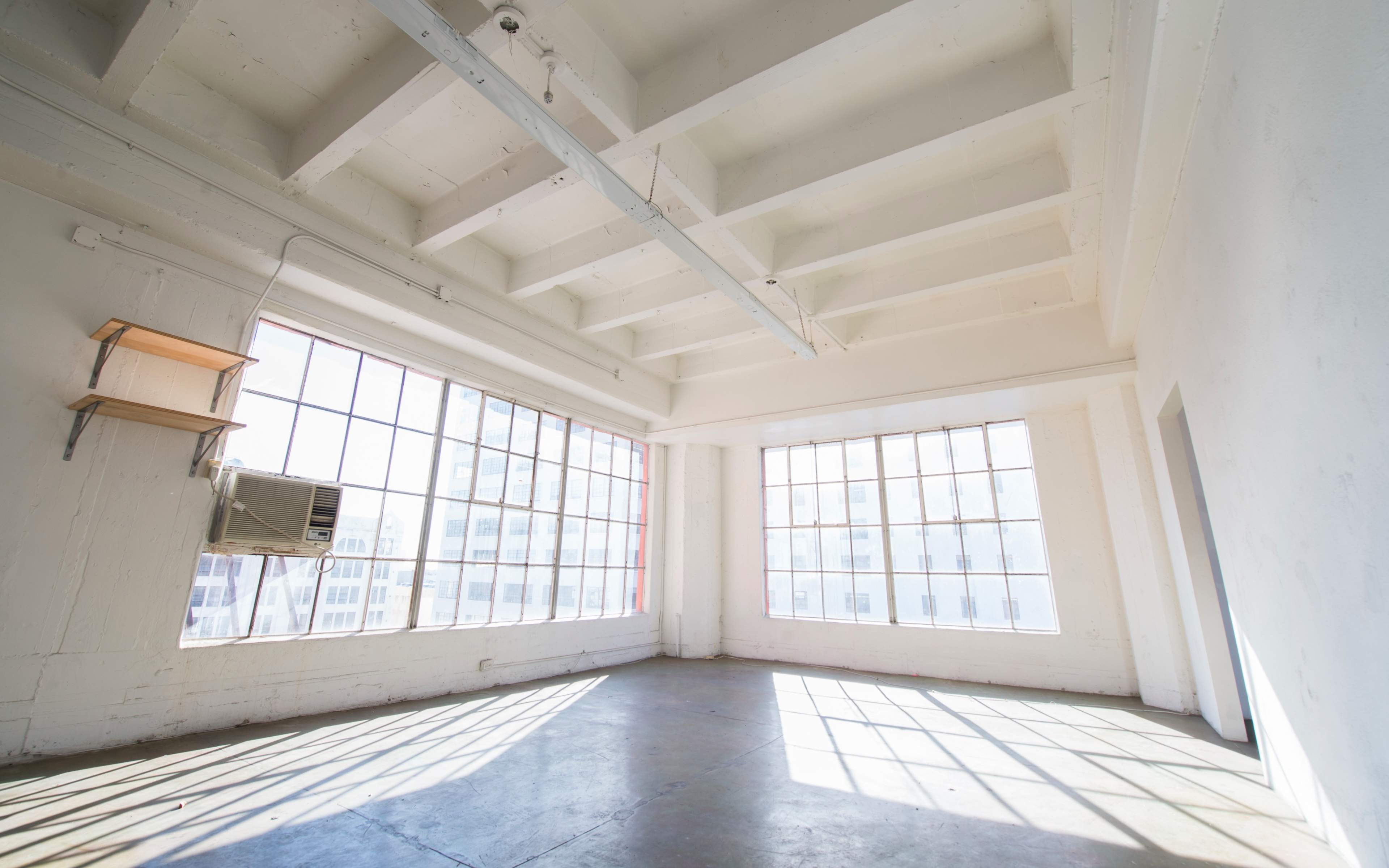 An empty room with large windows and a concrete floor, featuring exposed beams and a wall-mounted air conditioning unit.