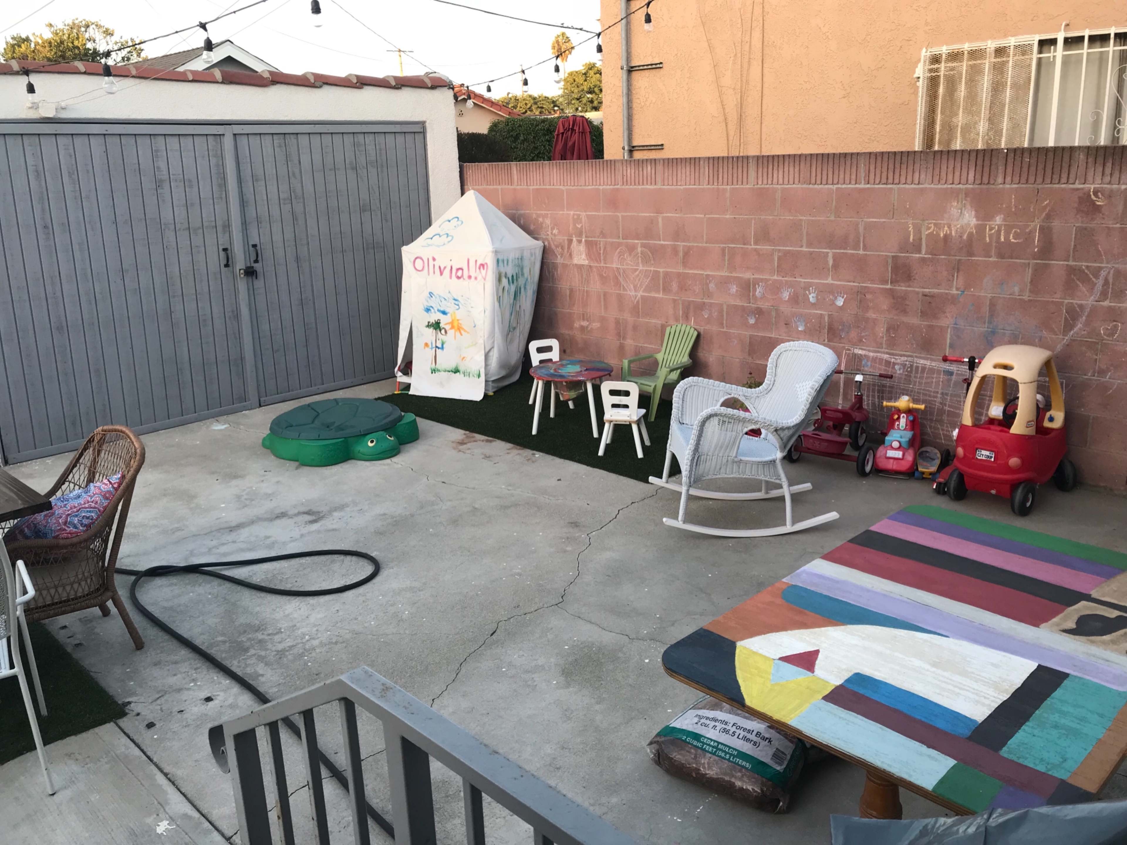 A concrete backyard area features a playhouse, colorful toys, outdoor furniture, and artificial grass.