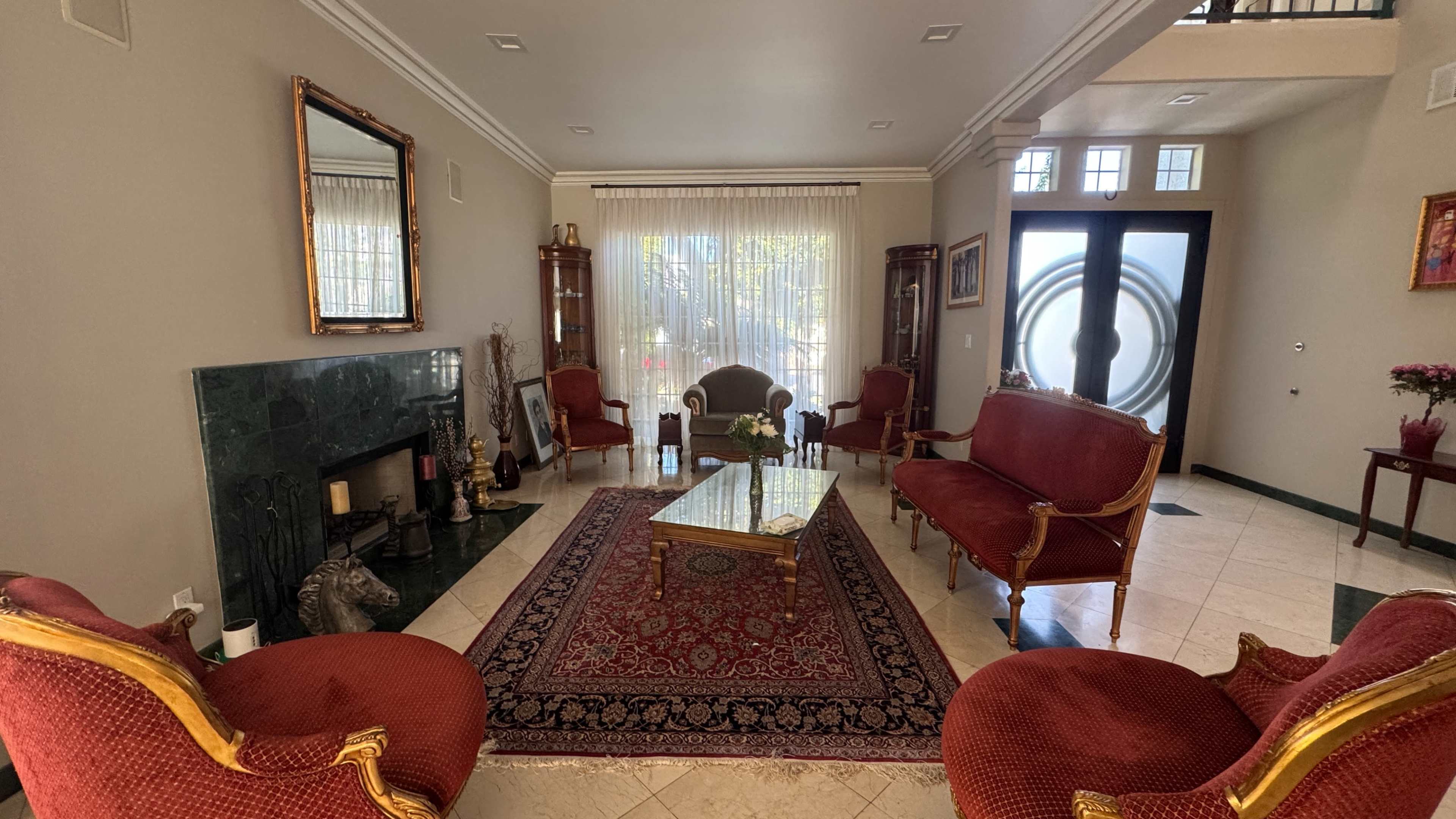 The image shows a spacious living room with a fireplace, ornate red chairs, a patterned rug, and large windows allowing natural light to enter.