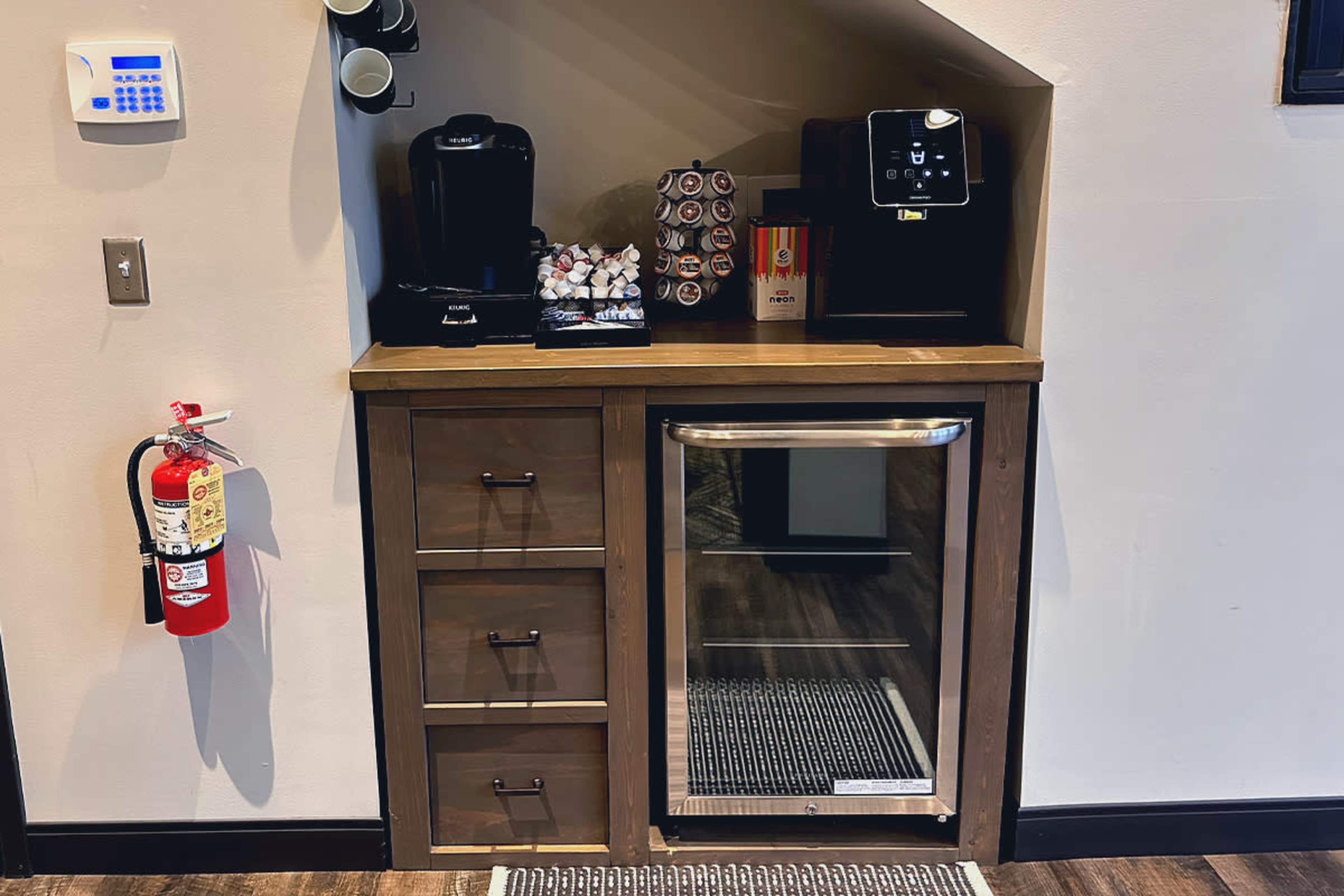 A kitchenette area featuring a coffee machine, a mini refrigerator, cabinets with three drawers, and containers of coffee pods on a wooden countertop.