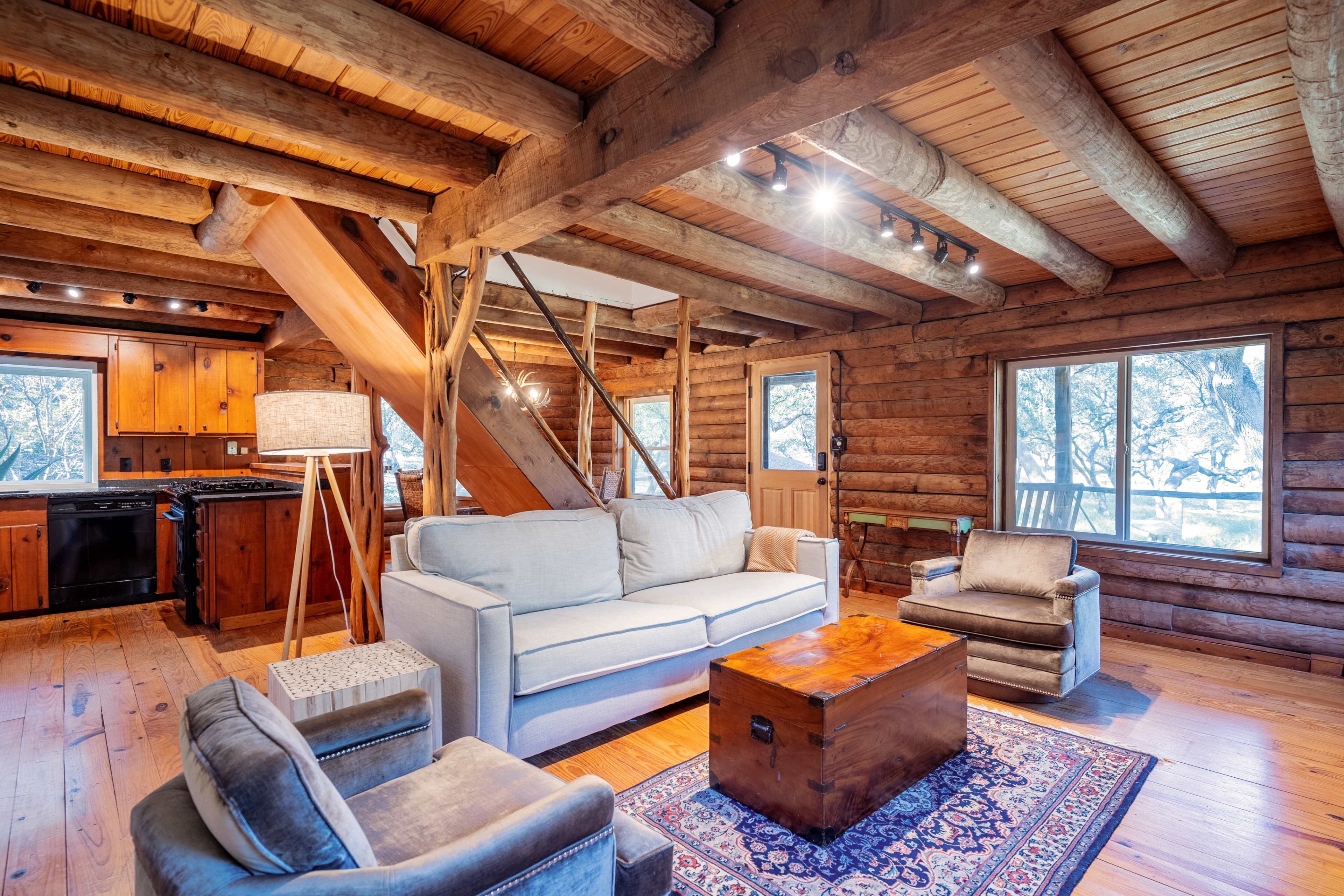 A rustic wooden cabin interior featuring a living area with a sofa, armchairs, a coffee table, and a kitchen area in the background.