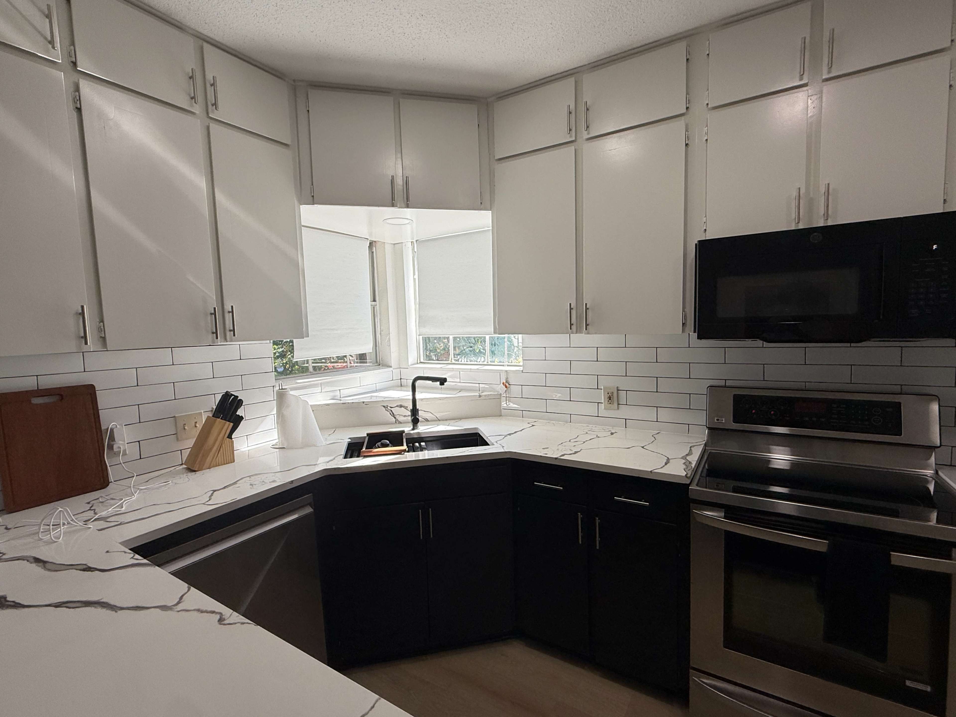 The image shows a modern kitchen featuring white cabinets, dark lower cabinets, a stainless steel microwave, and a marble-patterned countertop with a sink.