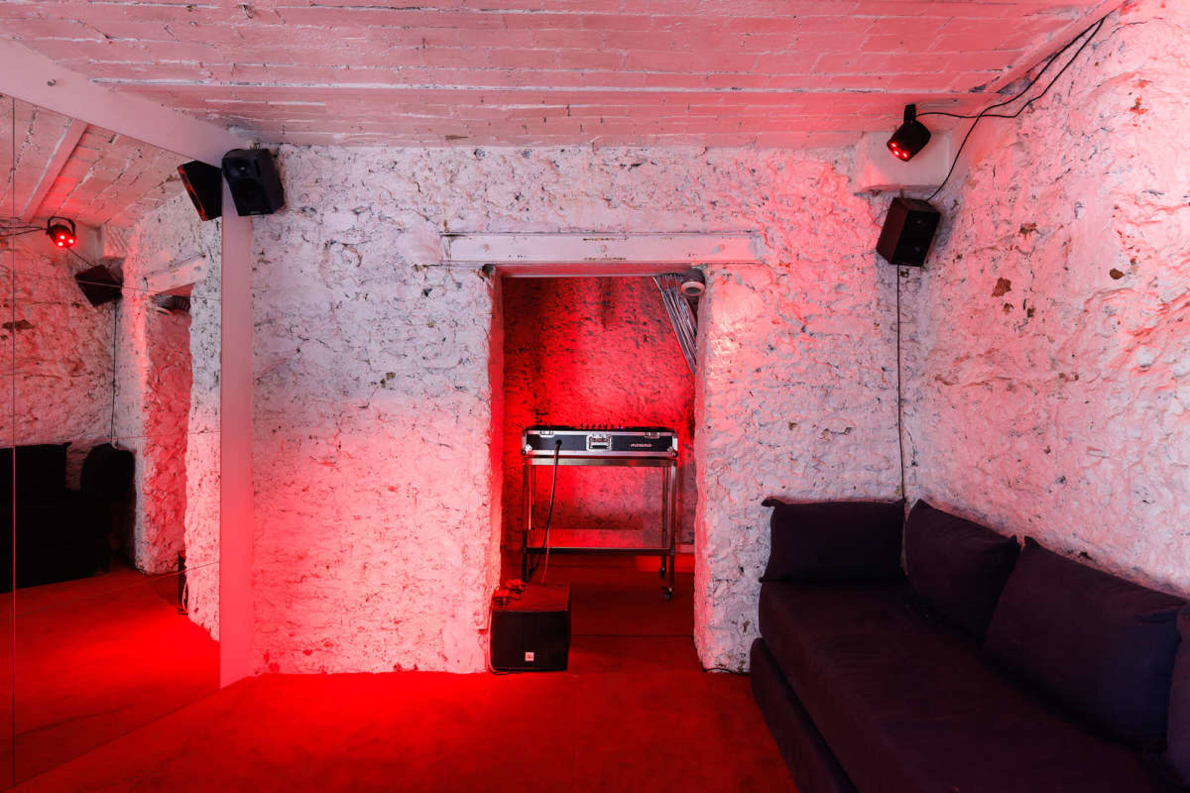 A dimly lit room features textured white walls, a red carpet, a black sofa, and a DJ setup in the background.