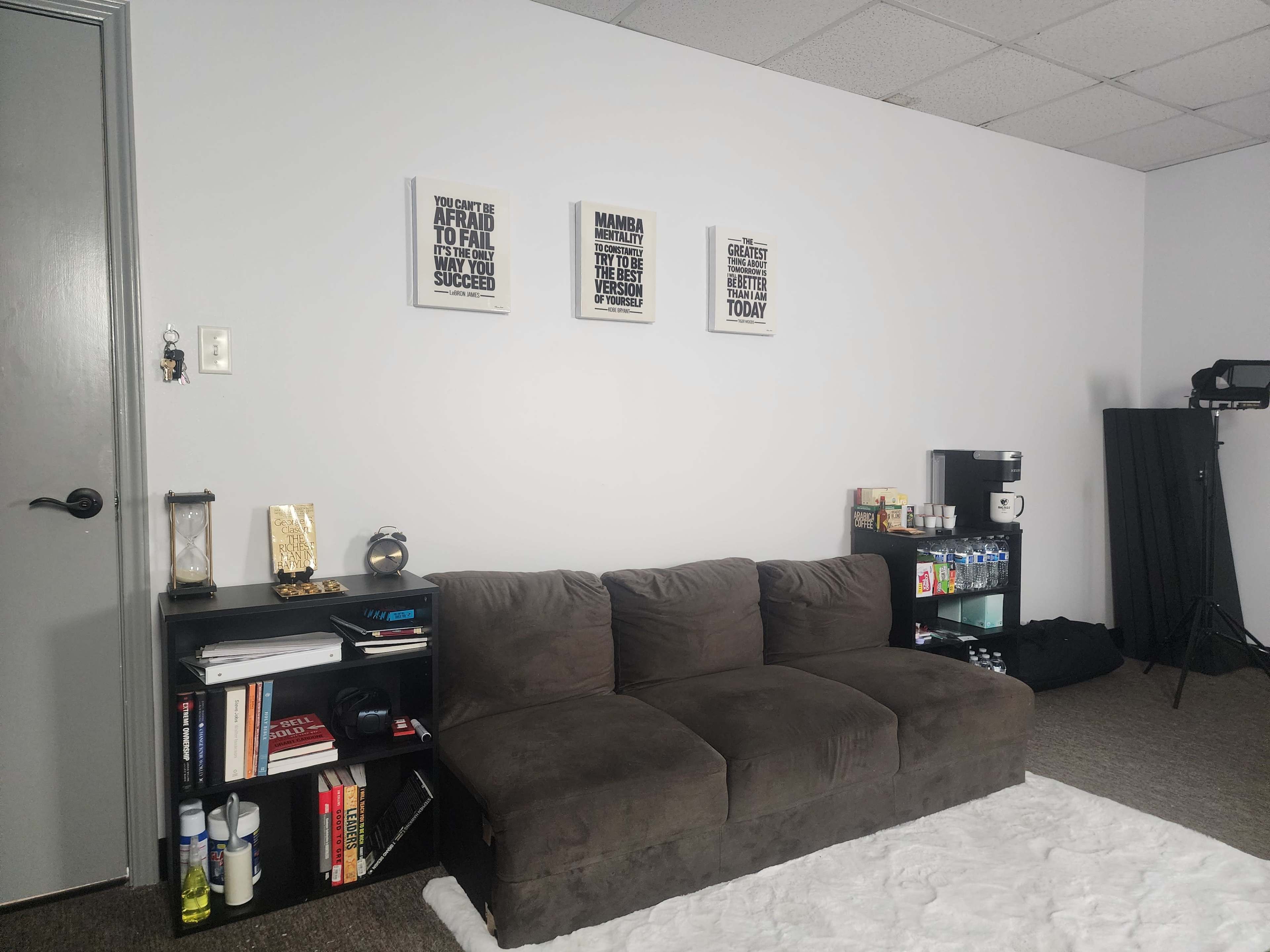 The image shows a cozy room featuring a brown couch against a light gray wall, adorned with motivational posters, and a shelf displaying books and various items beside a small refrigerator.