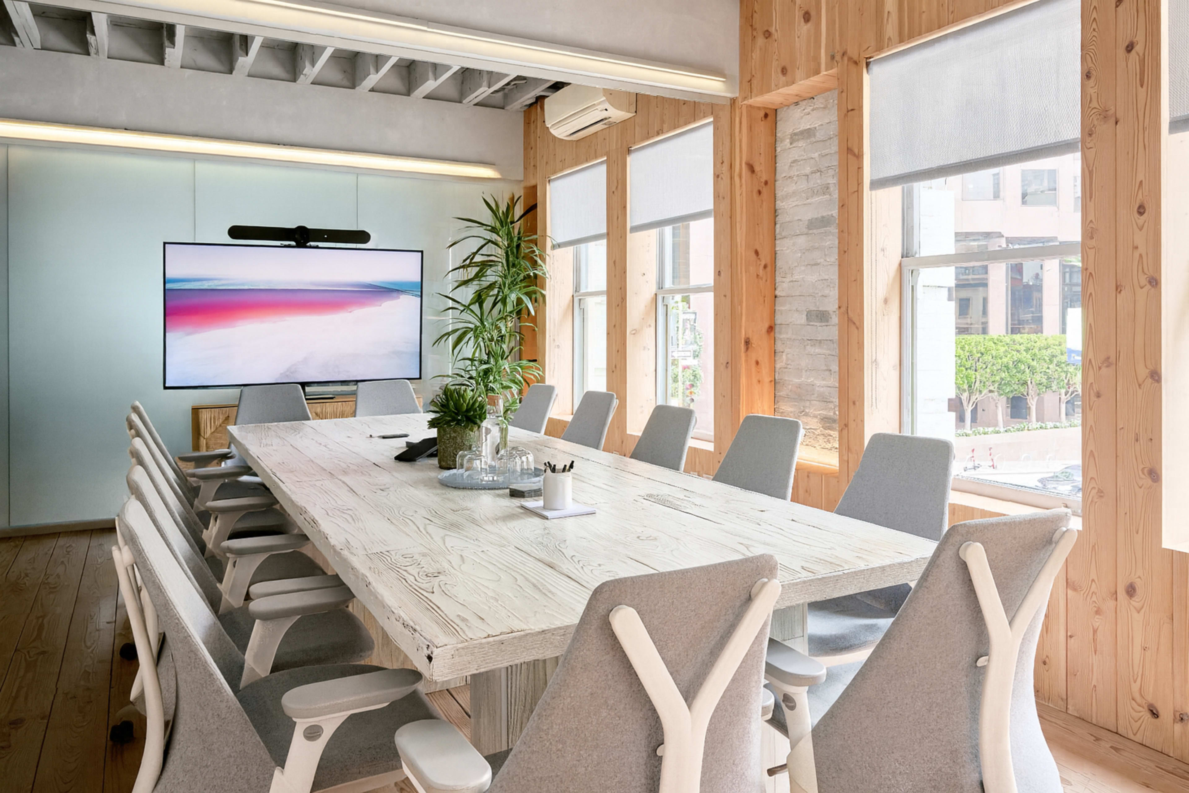 The image shows a modern conference room with a large wooden table, ergonomic chairs, and a screen displaying a landscape, illuminated by natural light through large windows.
